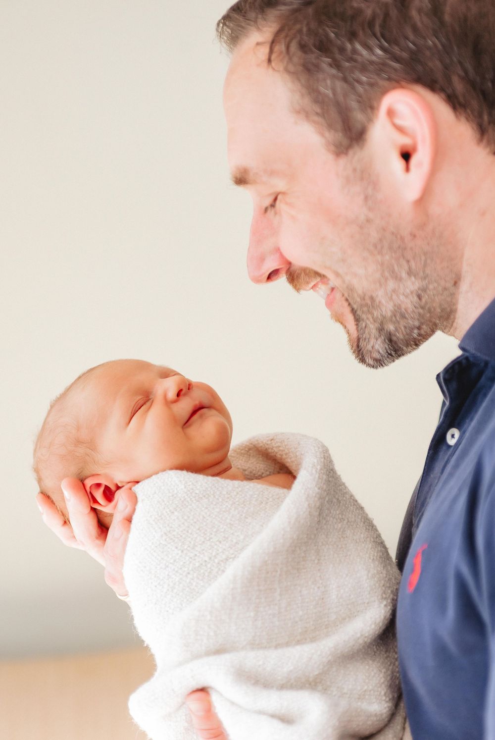 A smiling man with stubble holds a sleeping newborn baby wrapped in a white blanket, gazing down lovingly.