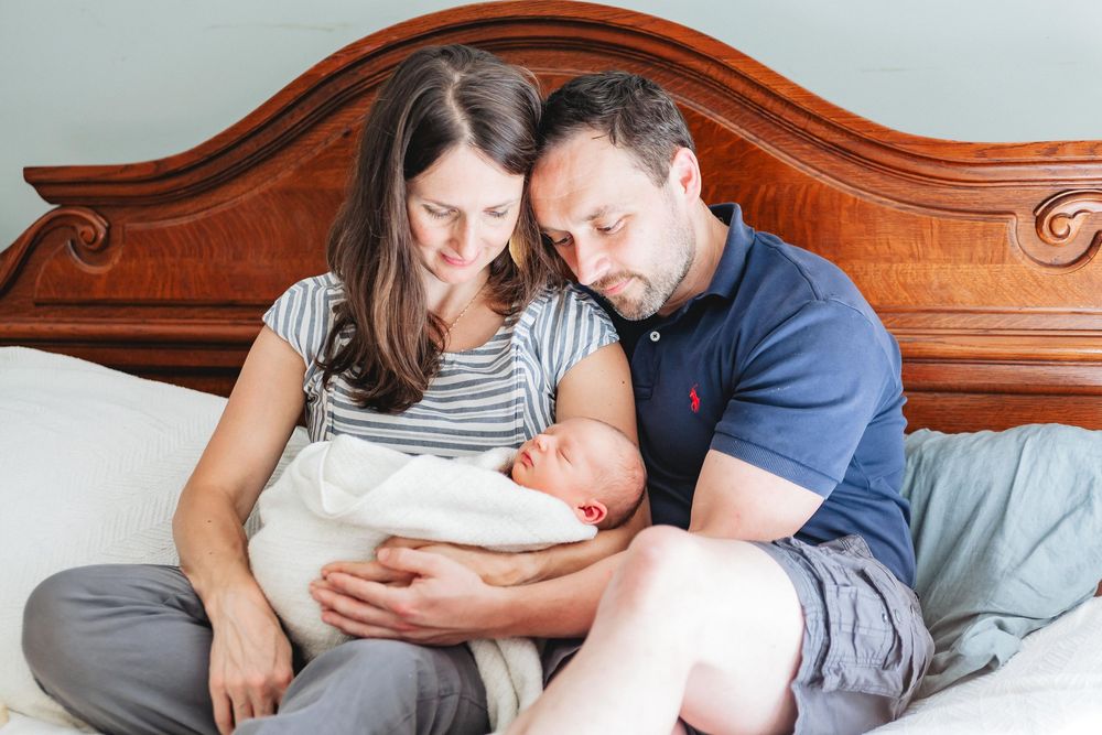 New parents sitting on a bed together, lovingly gazing at their swaddled newborn baby.