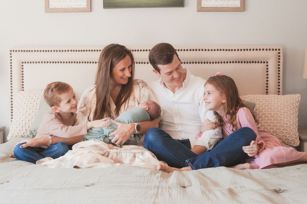 Happy family of five sitting on a bed, parents smiling at newborn baby with two young children beside them.