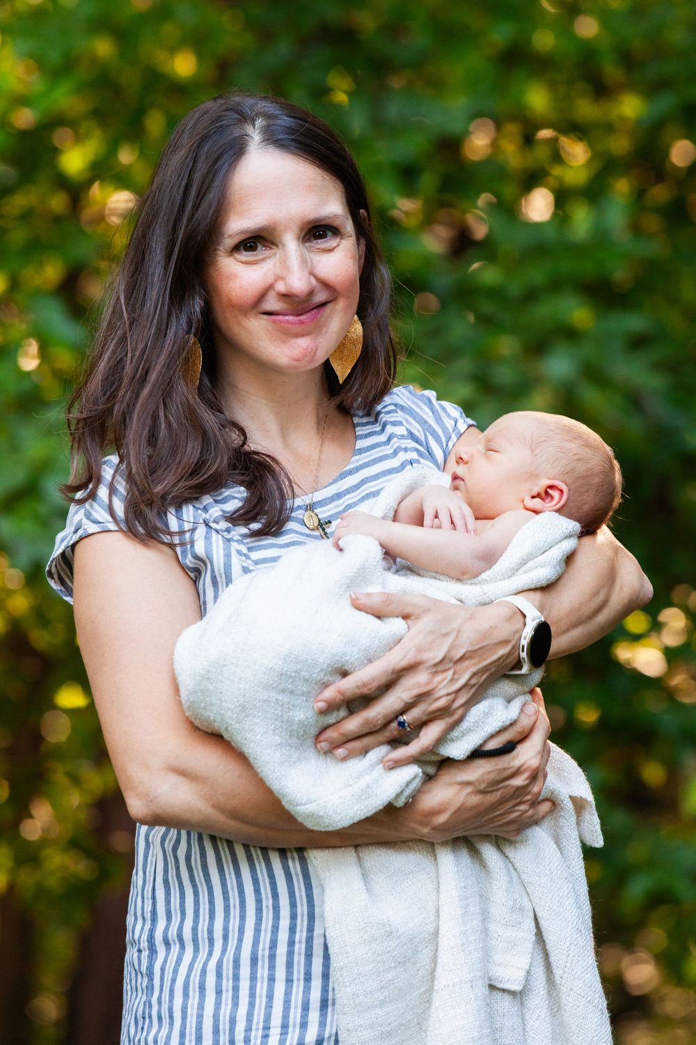 A smiling woman with dark hair holds a swaddled newborn baby outdoors with green trees in the background.