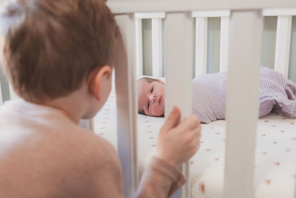 Older toddler peering through white crib rails at smiling newborn baby lying on dotted mattress.