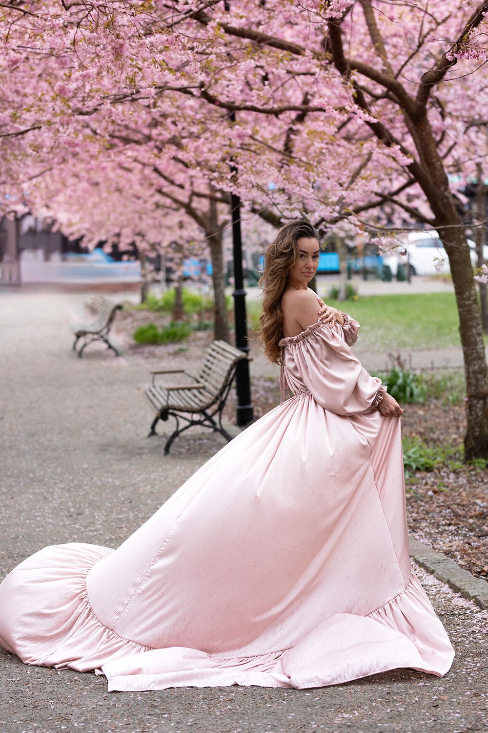 kvinna står i en park åt sidan med en rosa klänning. hon kollar in kameran och klänningen fylls av vind. i bakgrunden blommar körsbärsträd.