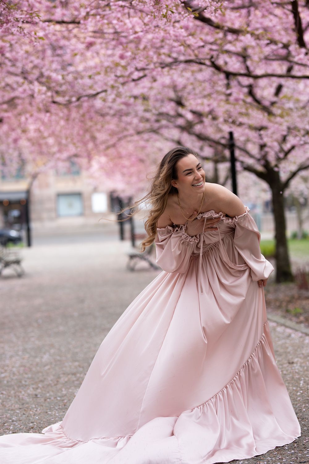 kvinna står i park och ler i en rosa klänning, i bakgrunden syns körsbärsträd blomma