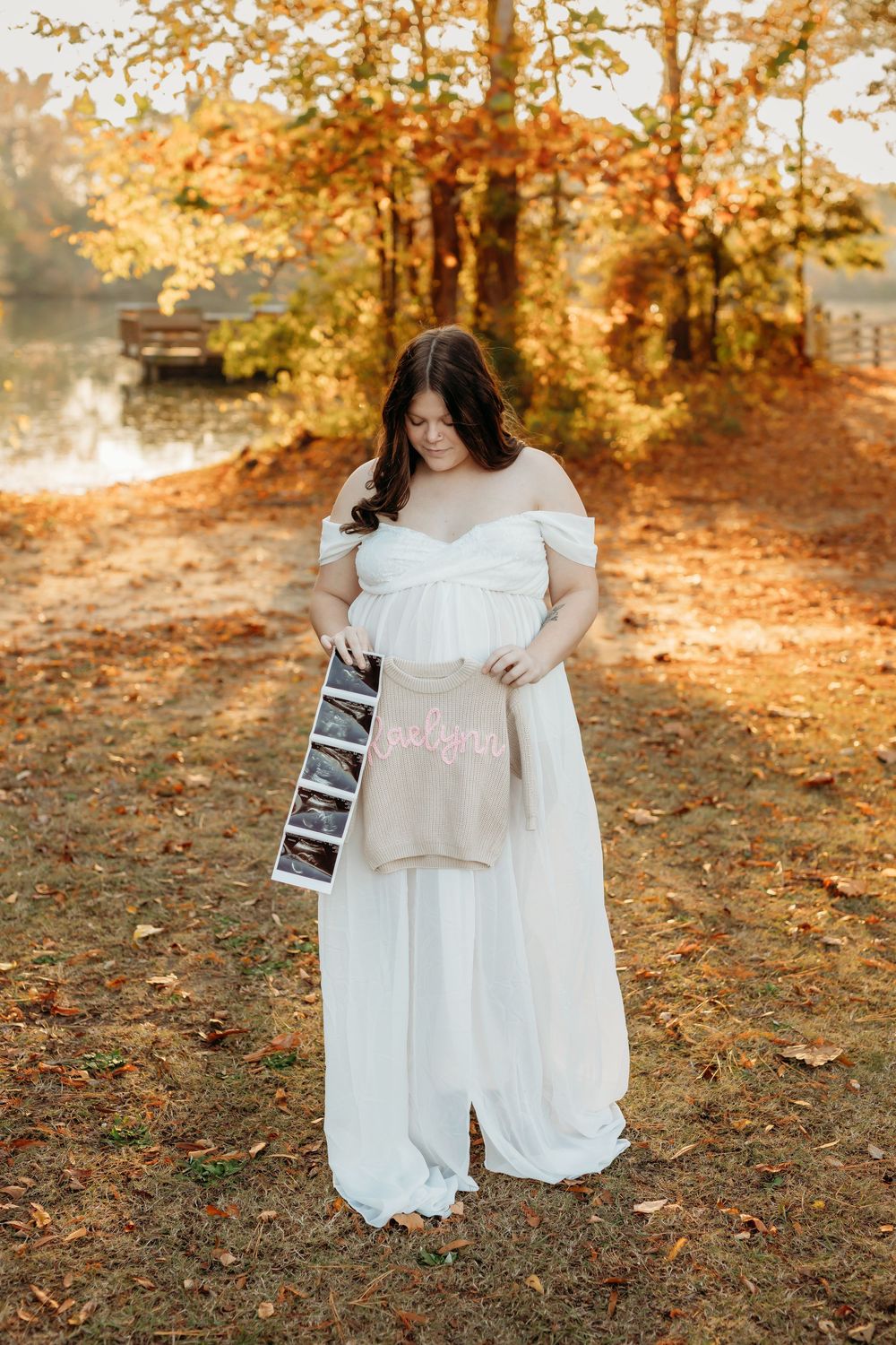 A maternity photoshoot in a white dress holding ultrasound photos during golden hour autumn.