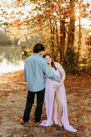 A romantic maternity photo of a couple embracing by a lake in autumn foliage.