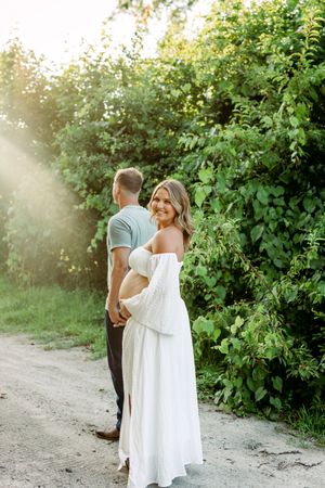 Maternity photo session on dirt path with natural sunlit greenery background.