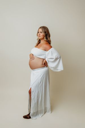 Solo maternity portraits in white dress against neutral backdrop.