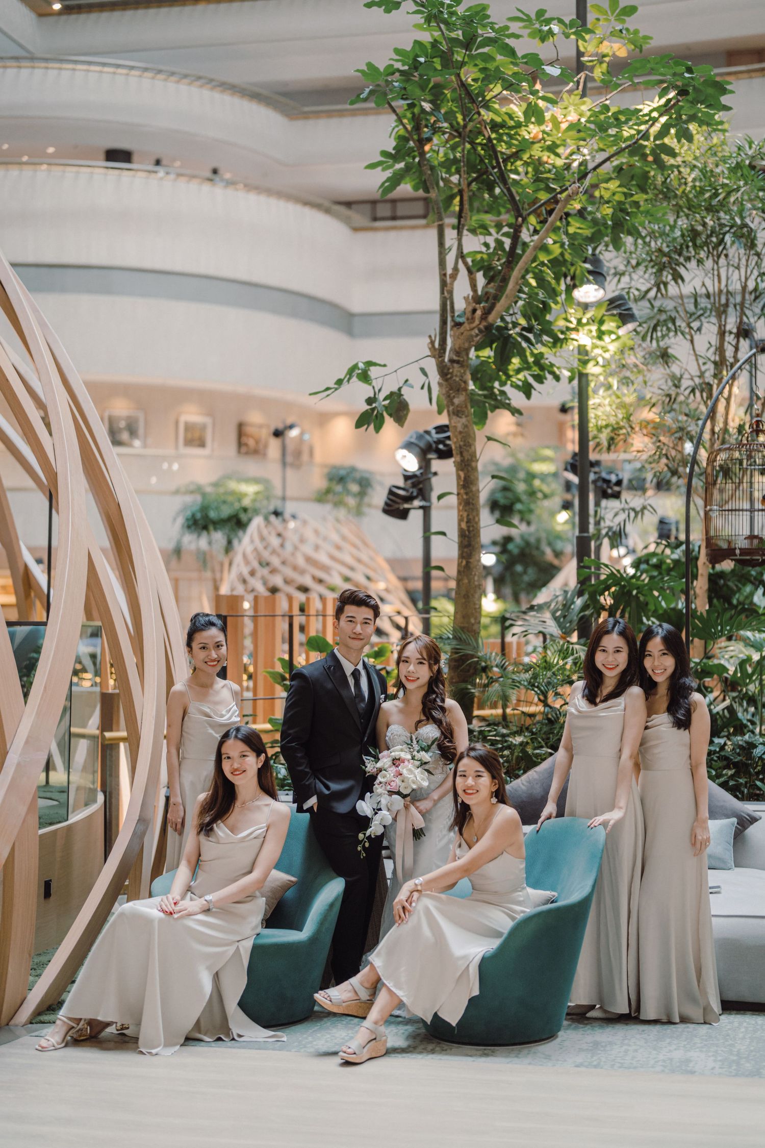 Wedding party relaxing on teal chairs in modern indoor garden setting with architectural details.