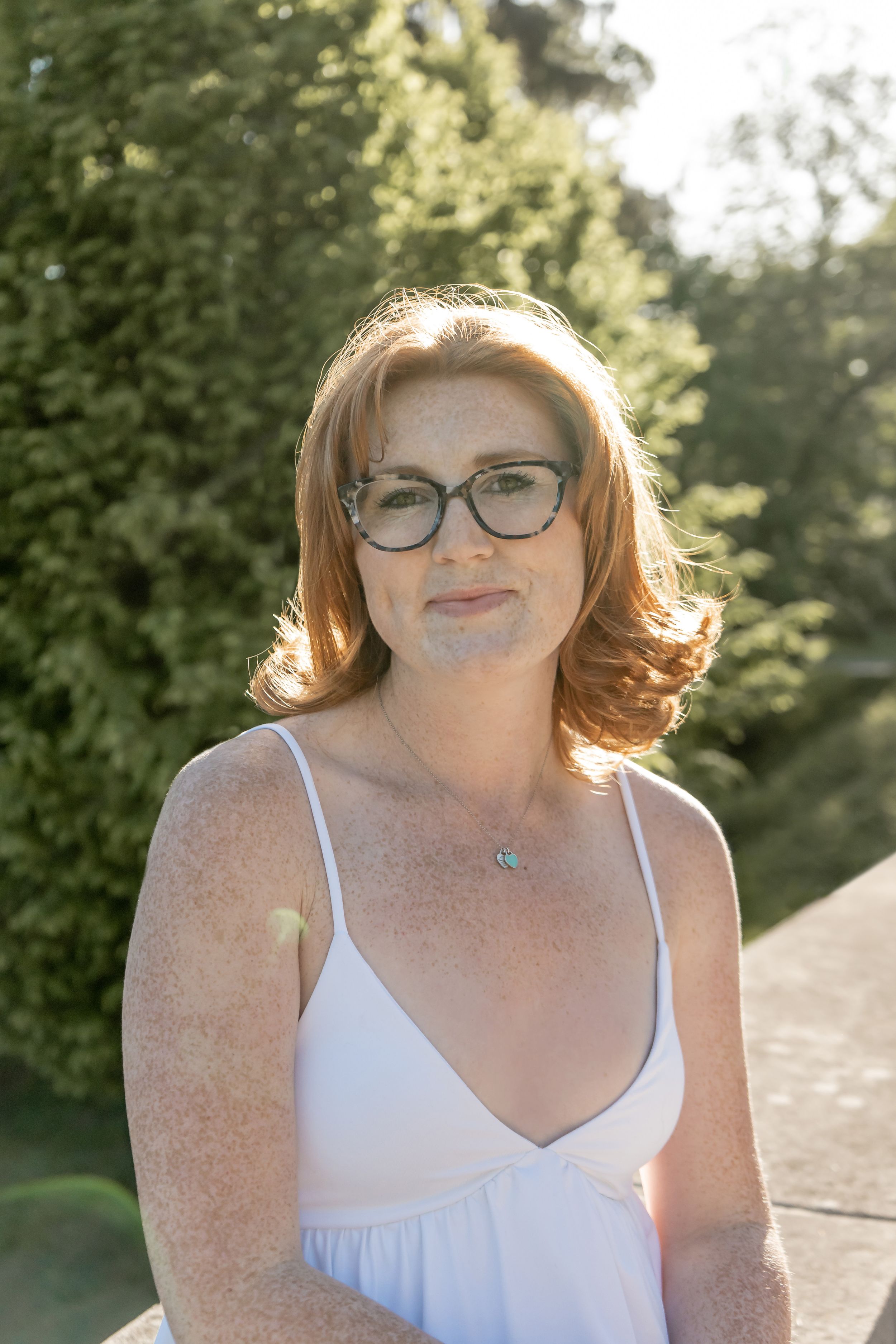 Red-haired woman with freckles and glasses posing outdoors in a white dress in warm sunlight.