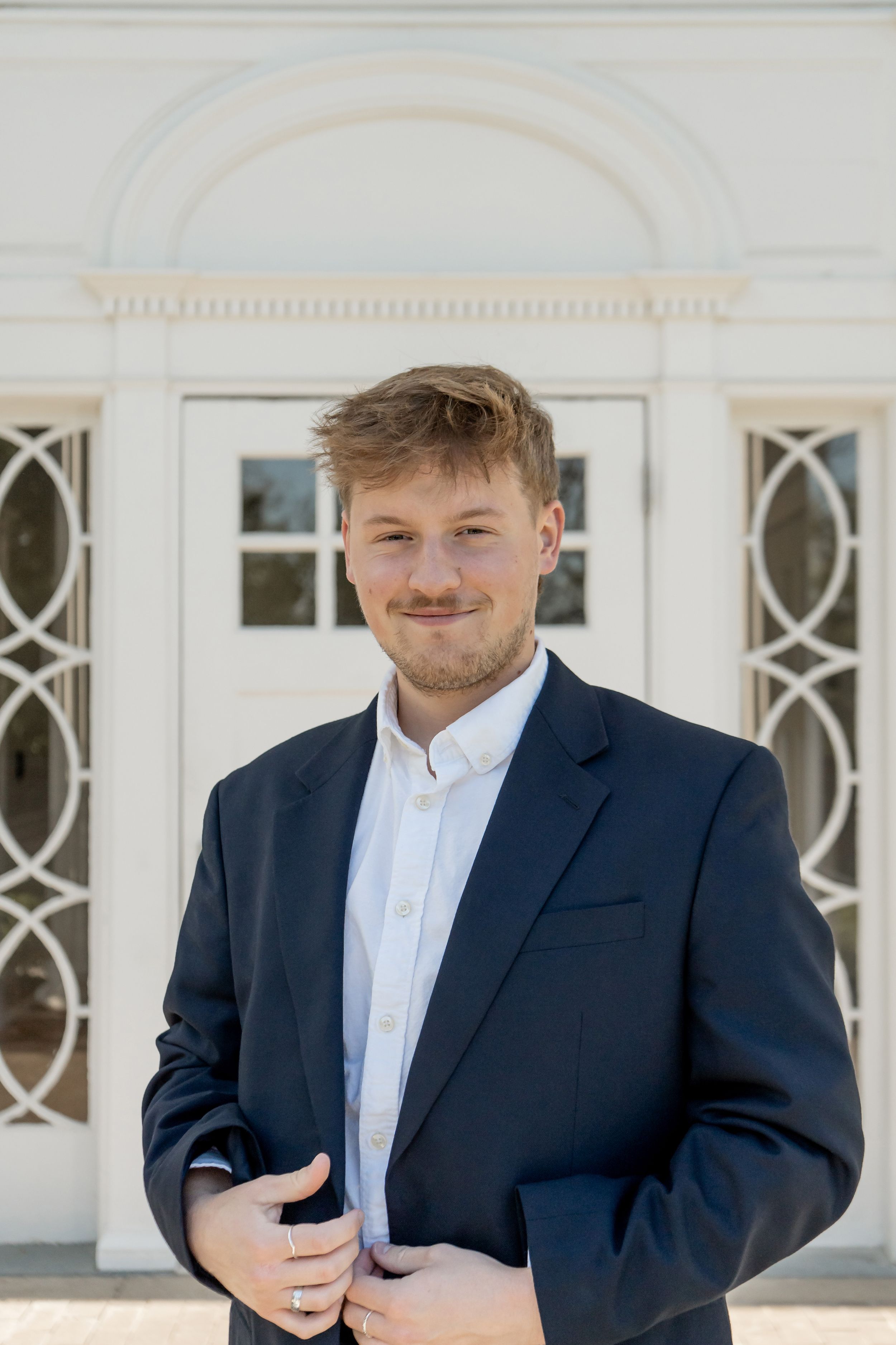 Young man in a navy blazer smiling confidently in front of a white decorative doorway outdoors.