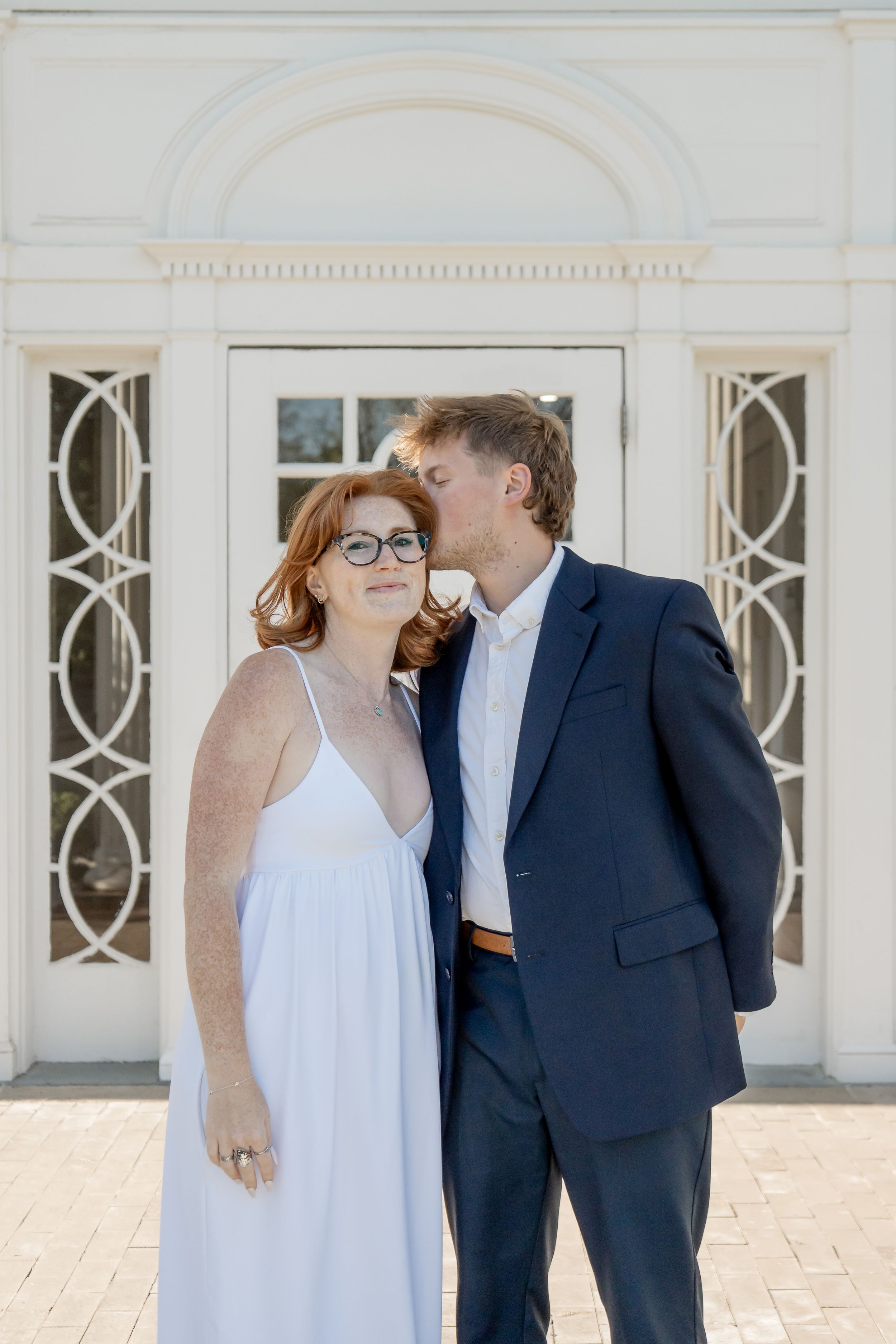 Couple posing in front of a white building, man in navy suit kissing woman in white dress.