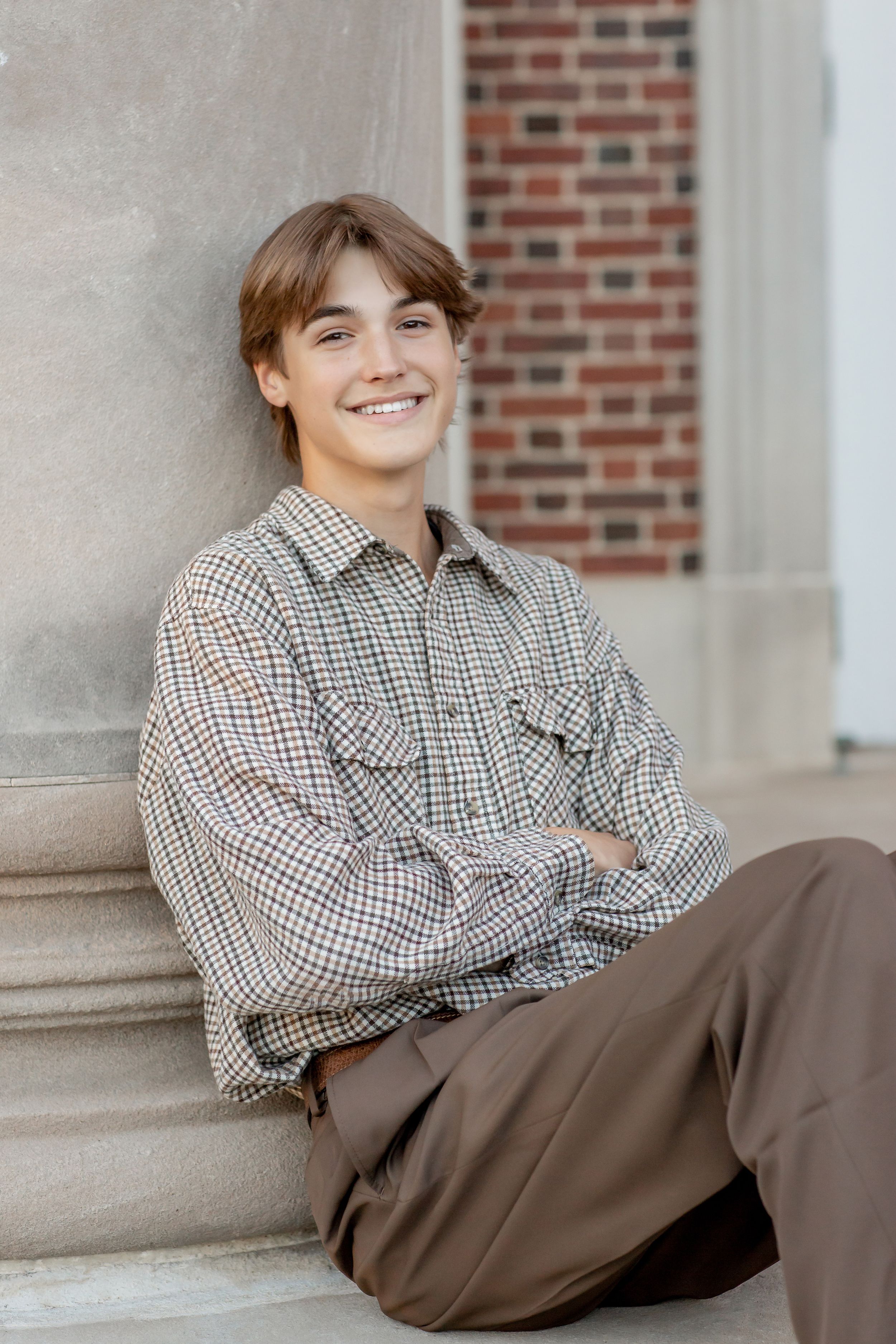 Smiling teenage boy in plaid shirt leaning against stone column outdoors.
