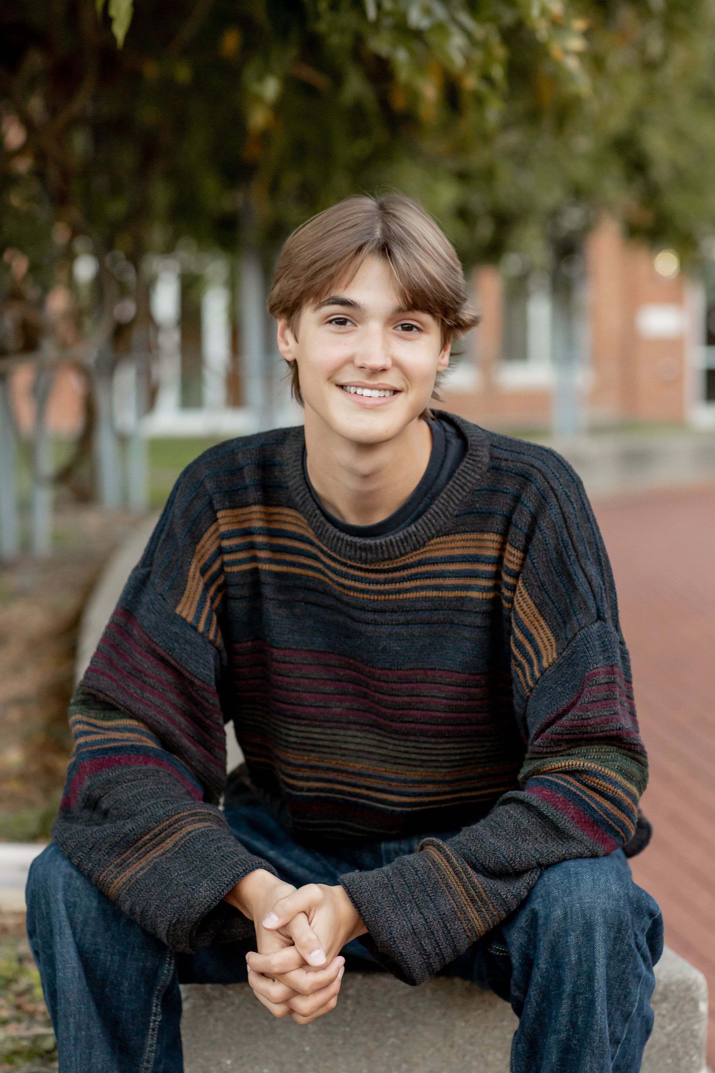 Smiling teen boy in striped sweater sitting on stone ledge in outdoor campus setting.