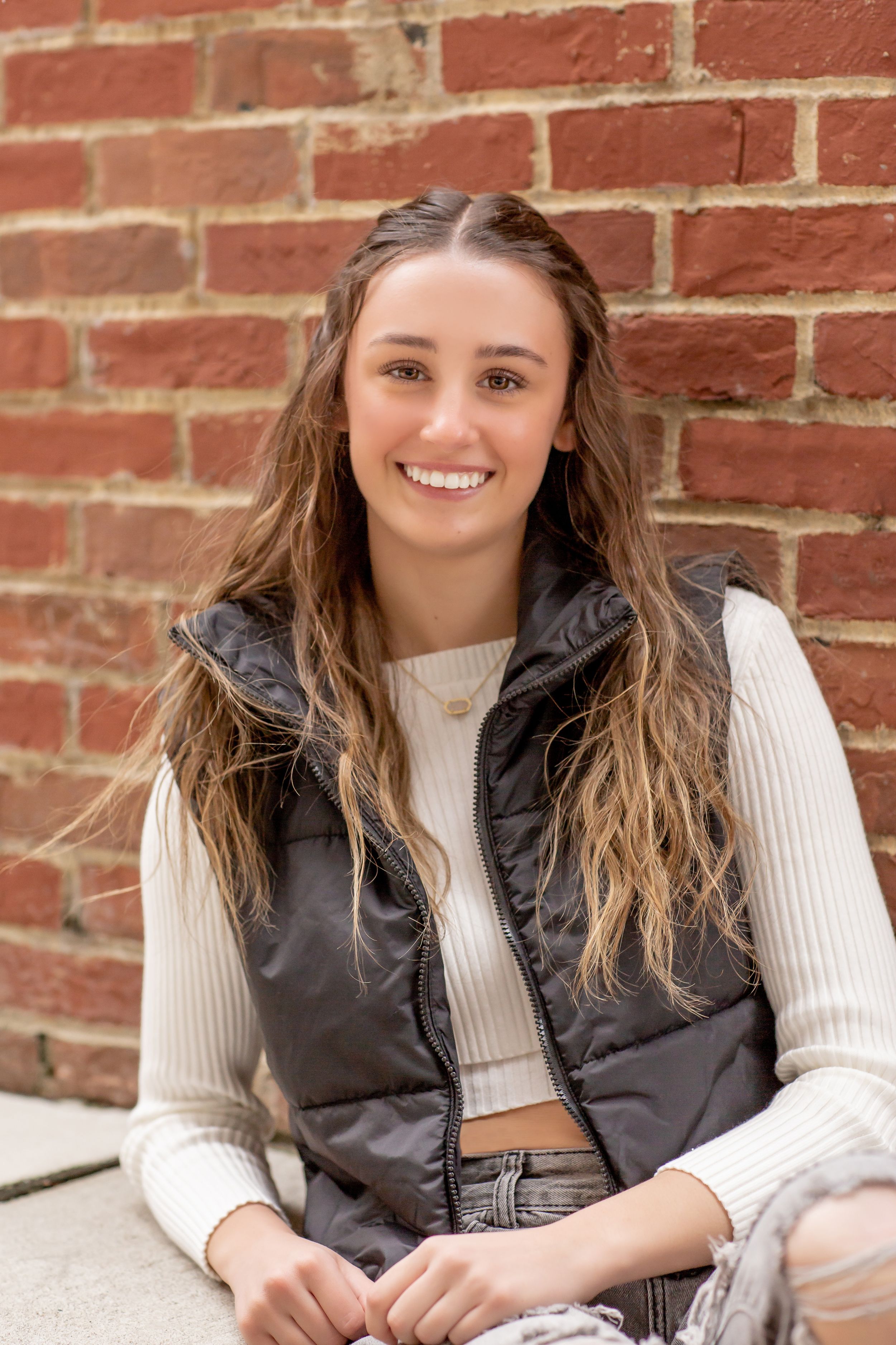 Smiling teenage girl with long wavy hair wearing a black puffer vest sitting against a red brick wall.