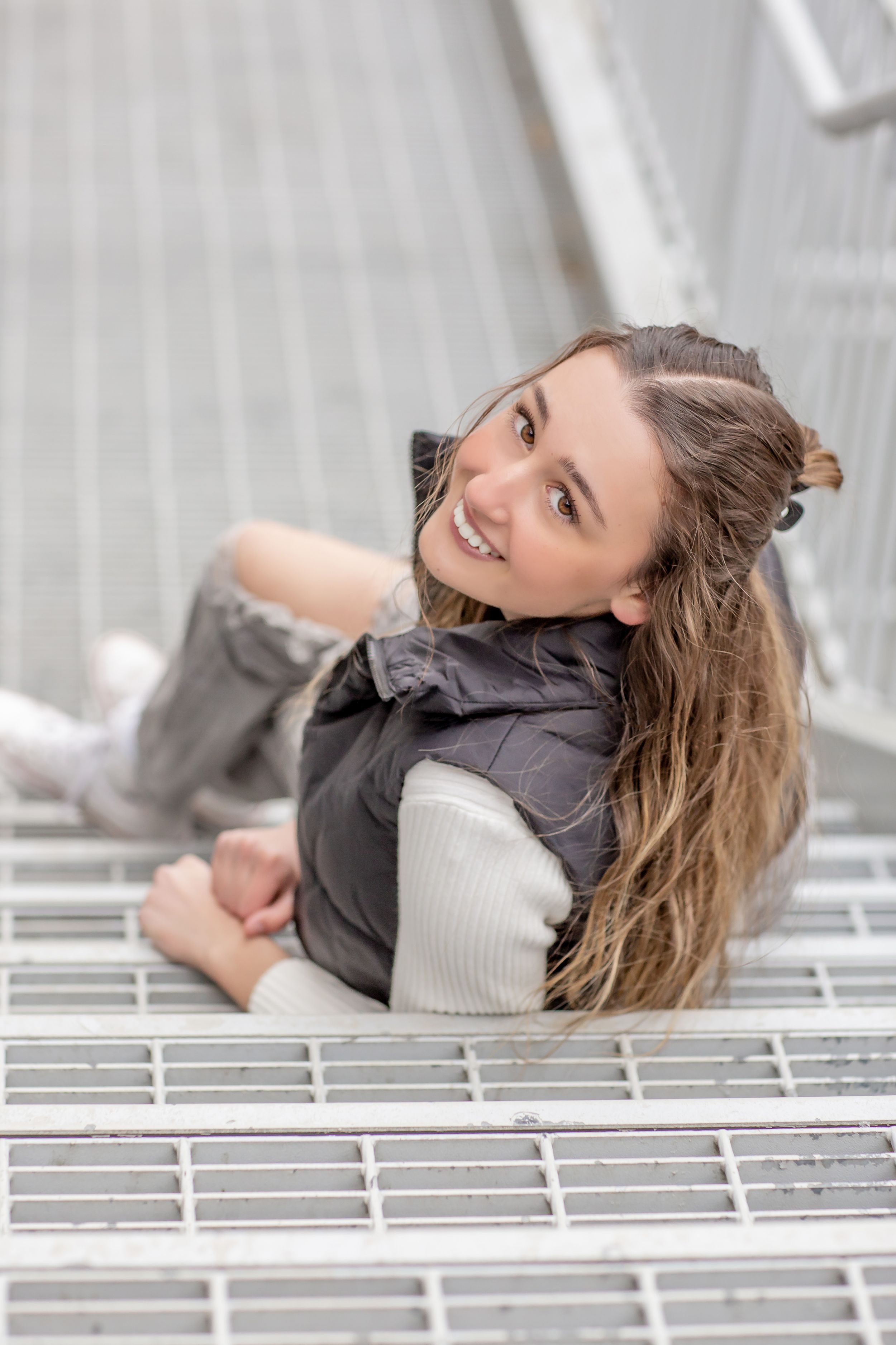 Smiling teenage girl with long brown hair sitting on metal grate stairs wearing a gray vest and sweater.