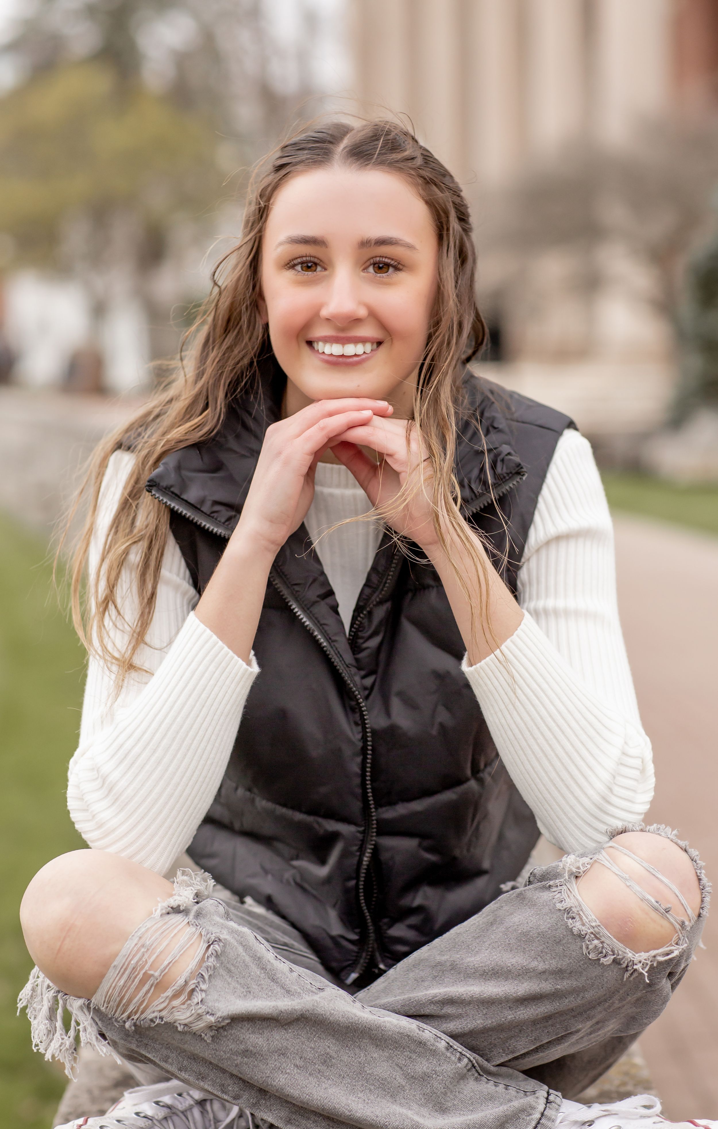 Smiling teen girl with long hair wearing a black vest and white sweater sitting cross-legged outdoors.