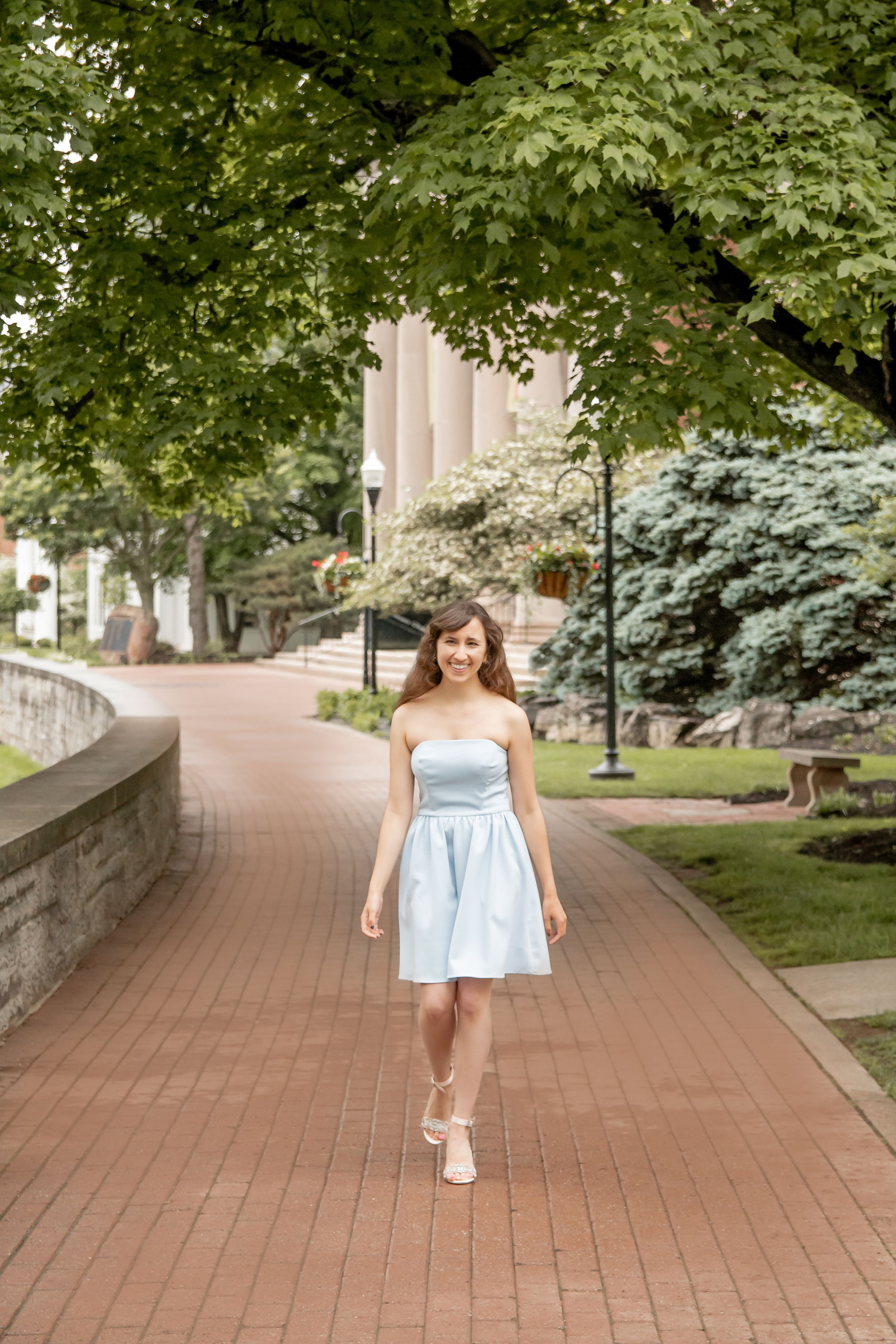 Smiling young woman in light blue strapless dress walking on brick campus pathway.