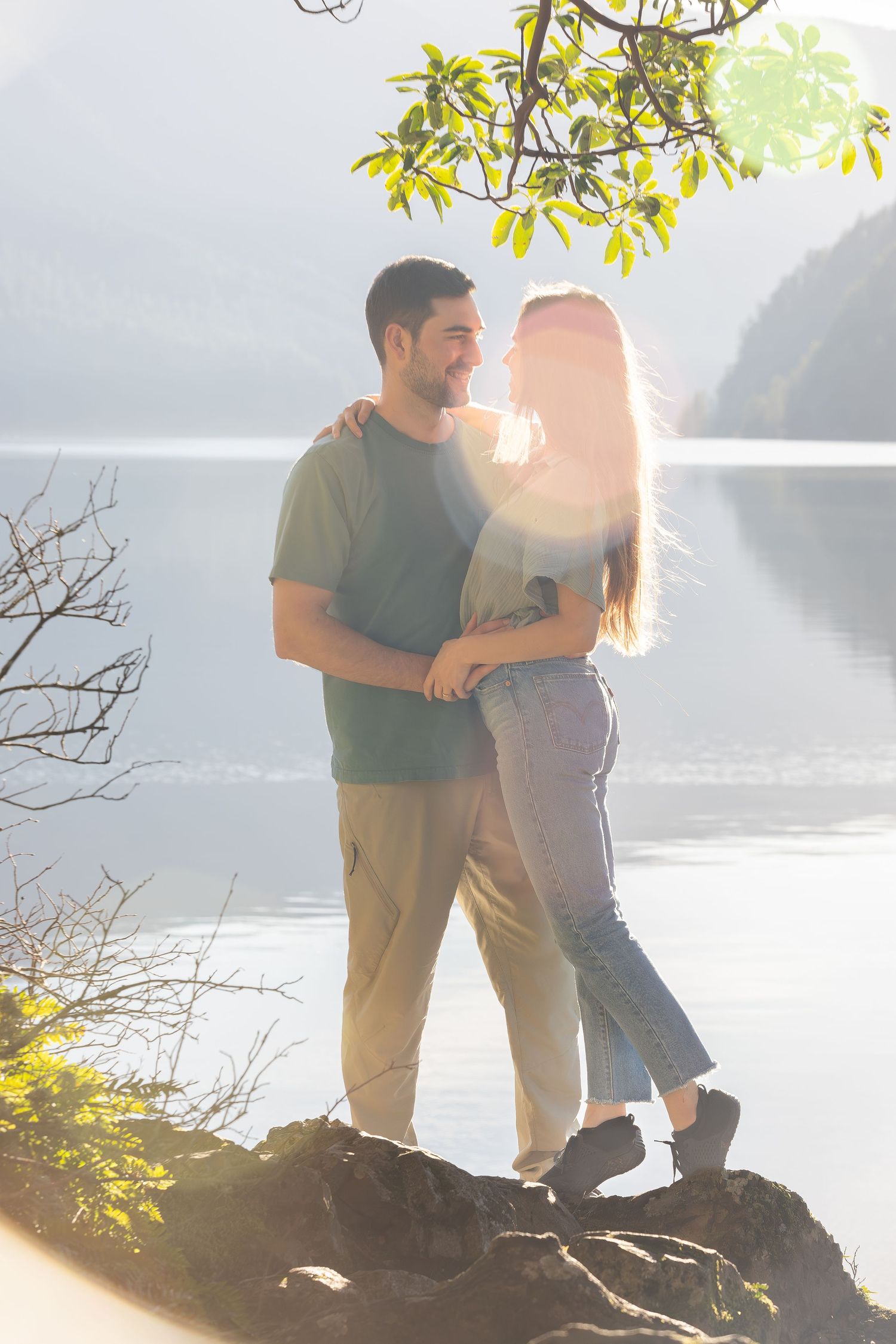 Couple sharing intimate moment by lakeside with sunlit mountain backdrop.