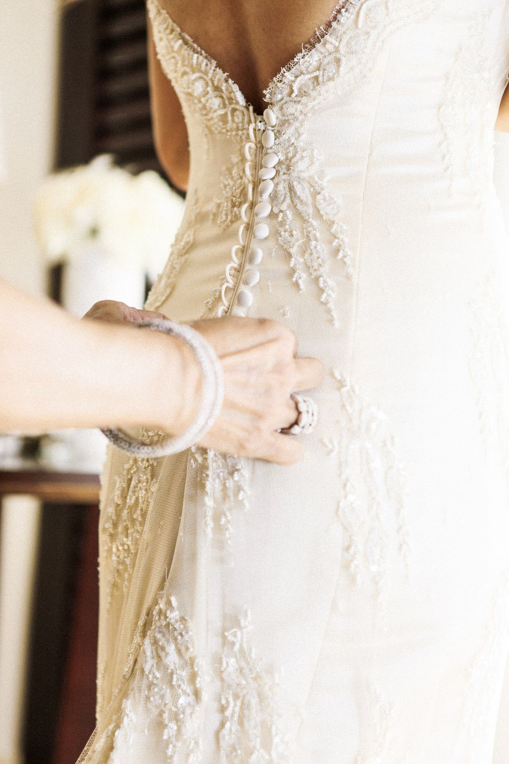 Close-up detail of a white lace wedding dress with delicate beading and a bracelet-adorned hand adjusting the gown.
