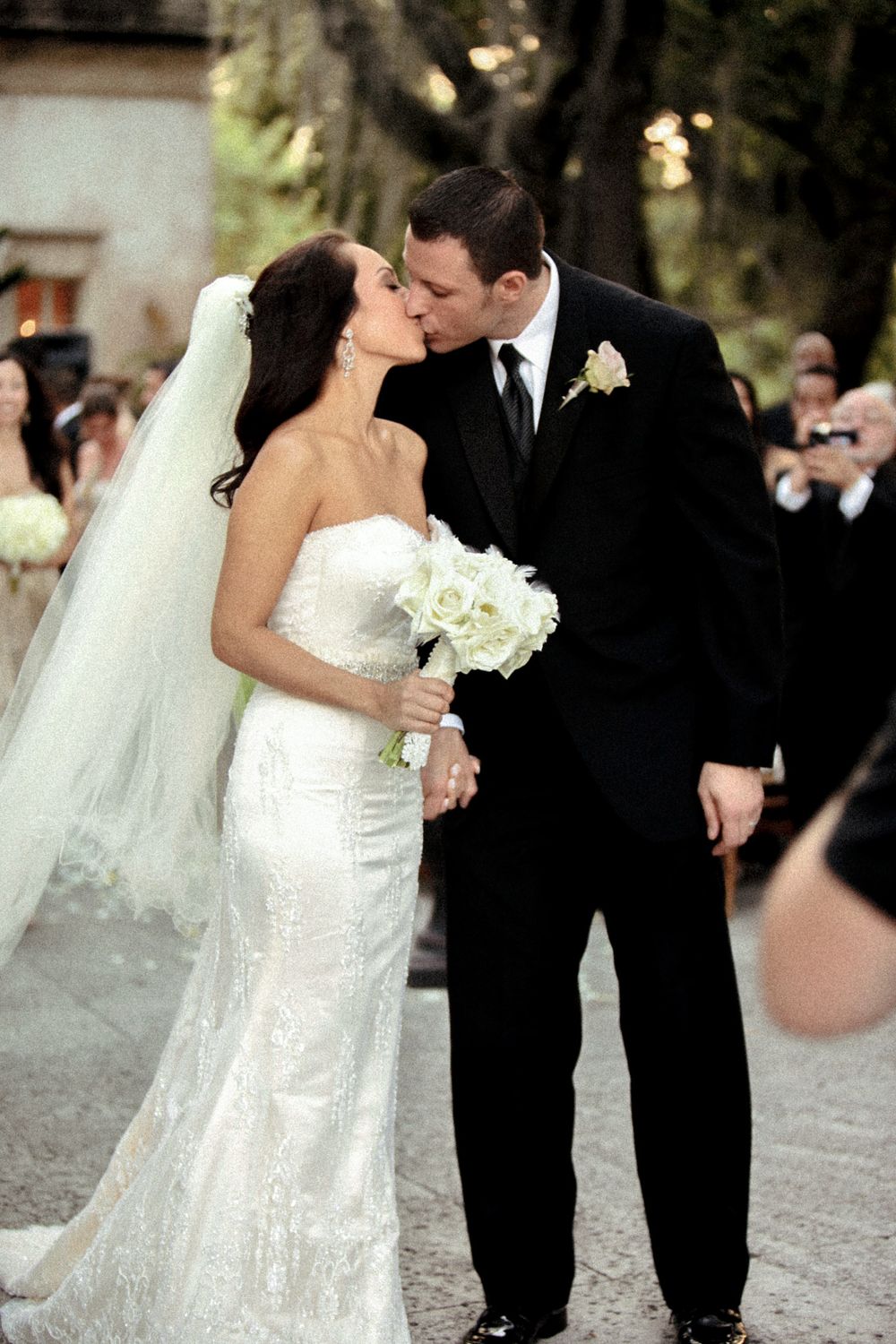 Wedding couple sharing a romantic kiss with guests in the background during an outdoor ceremony.