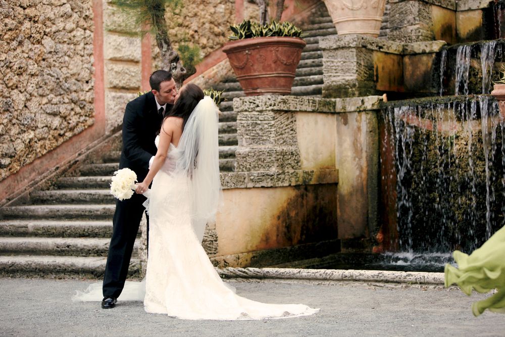 Wedding couple stands in an elegant pose near stone steps and a cascading fountain at a historic venue.