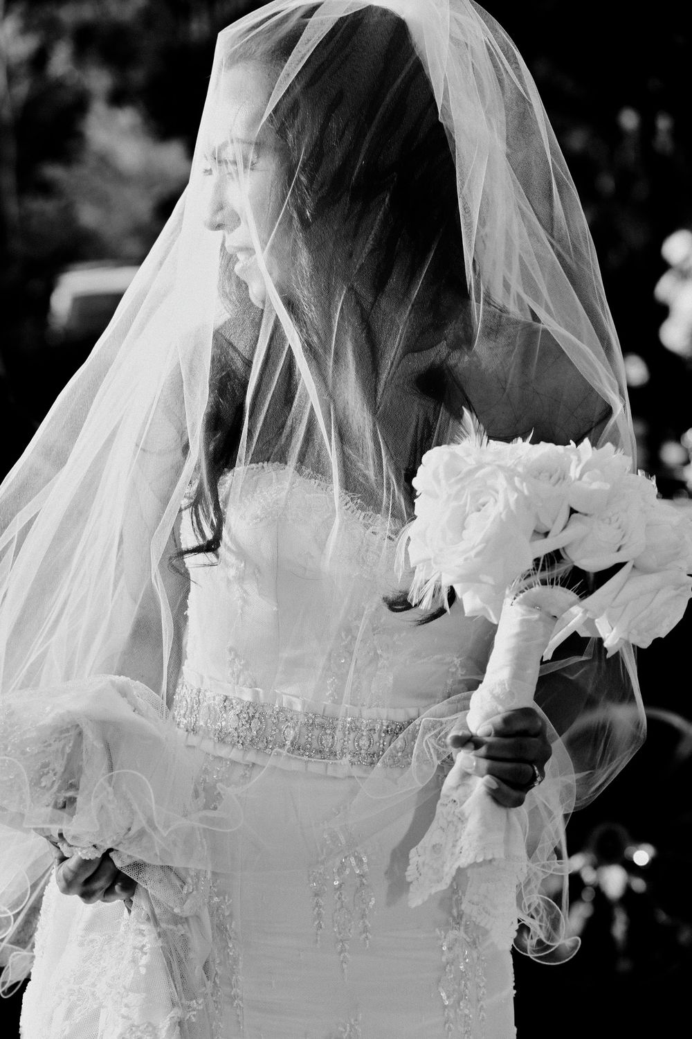 A bride in a white wedding gown and veil holds a bouquet of white flowers in this black and white photograph.