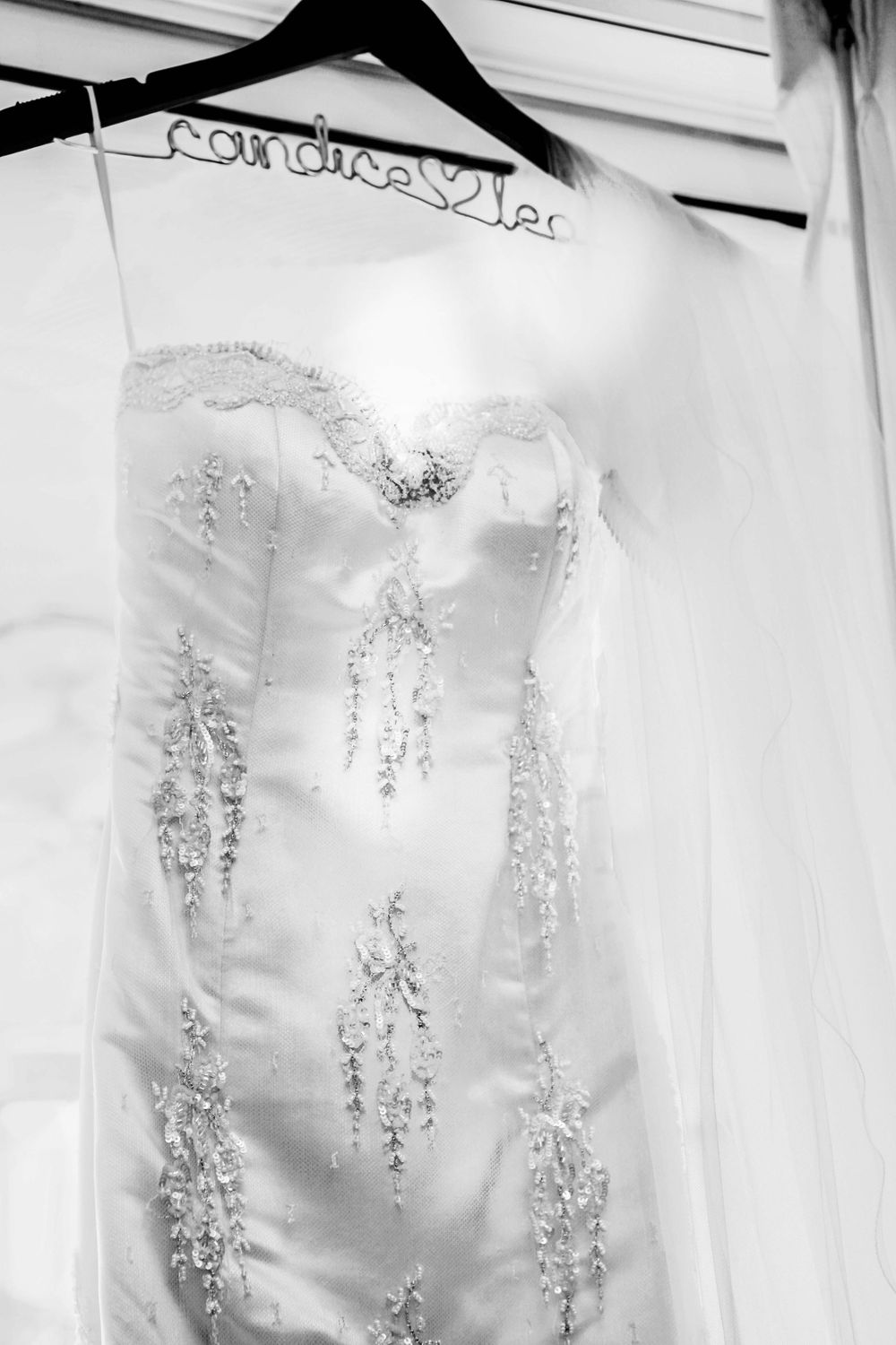 A white wedding dress with intricate beaded embellishments hangs on a decorative hanger in black and white photography.