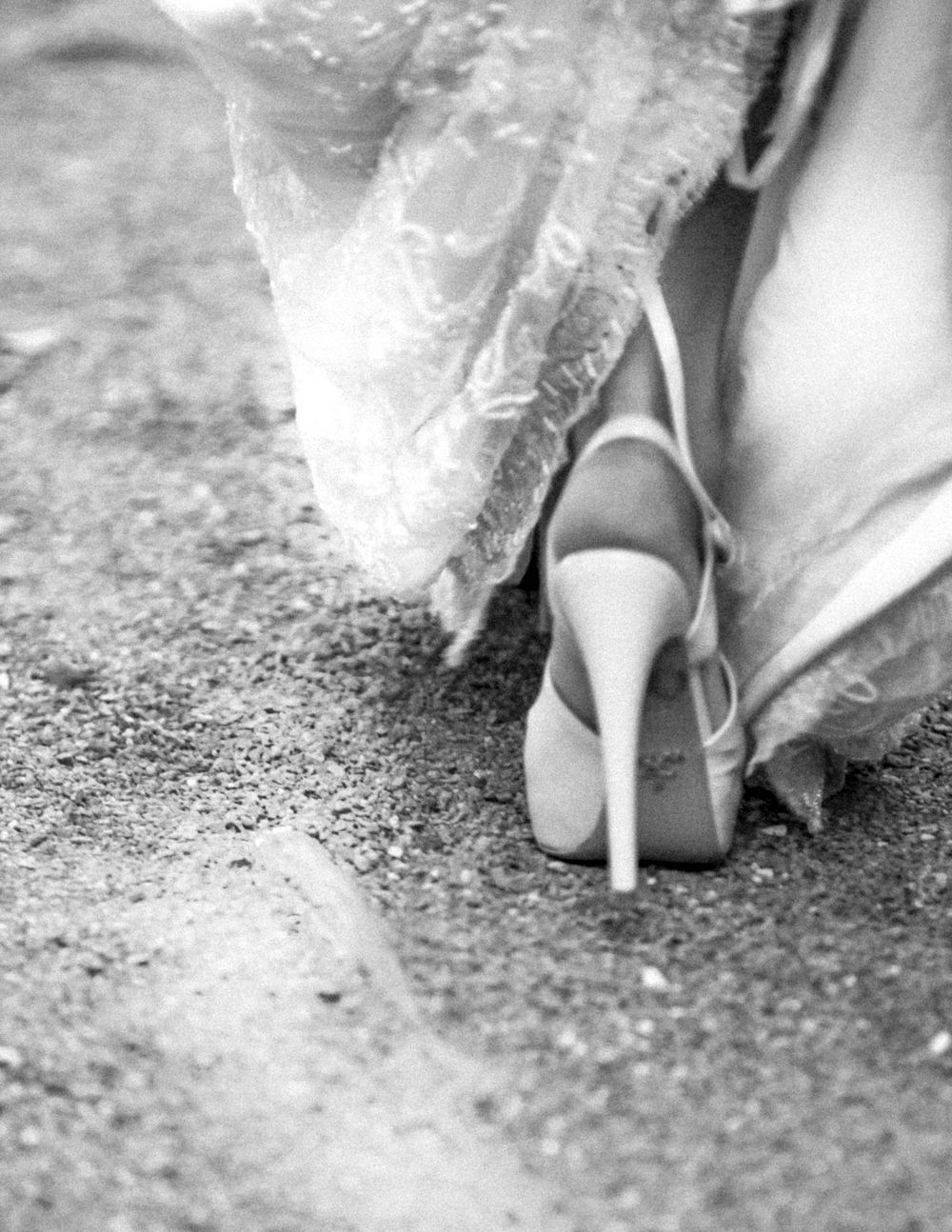 A close-up black and white photo shows an elegant high-heeled shoe peeking out from beneath a lace wedding dress hem.