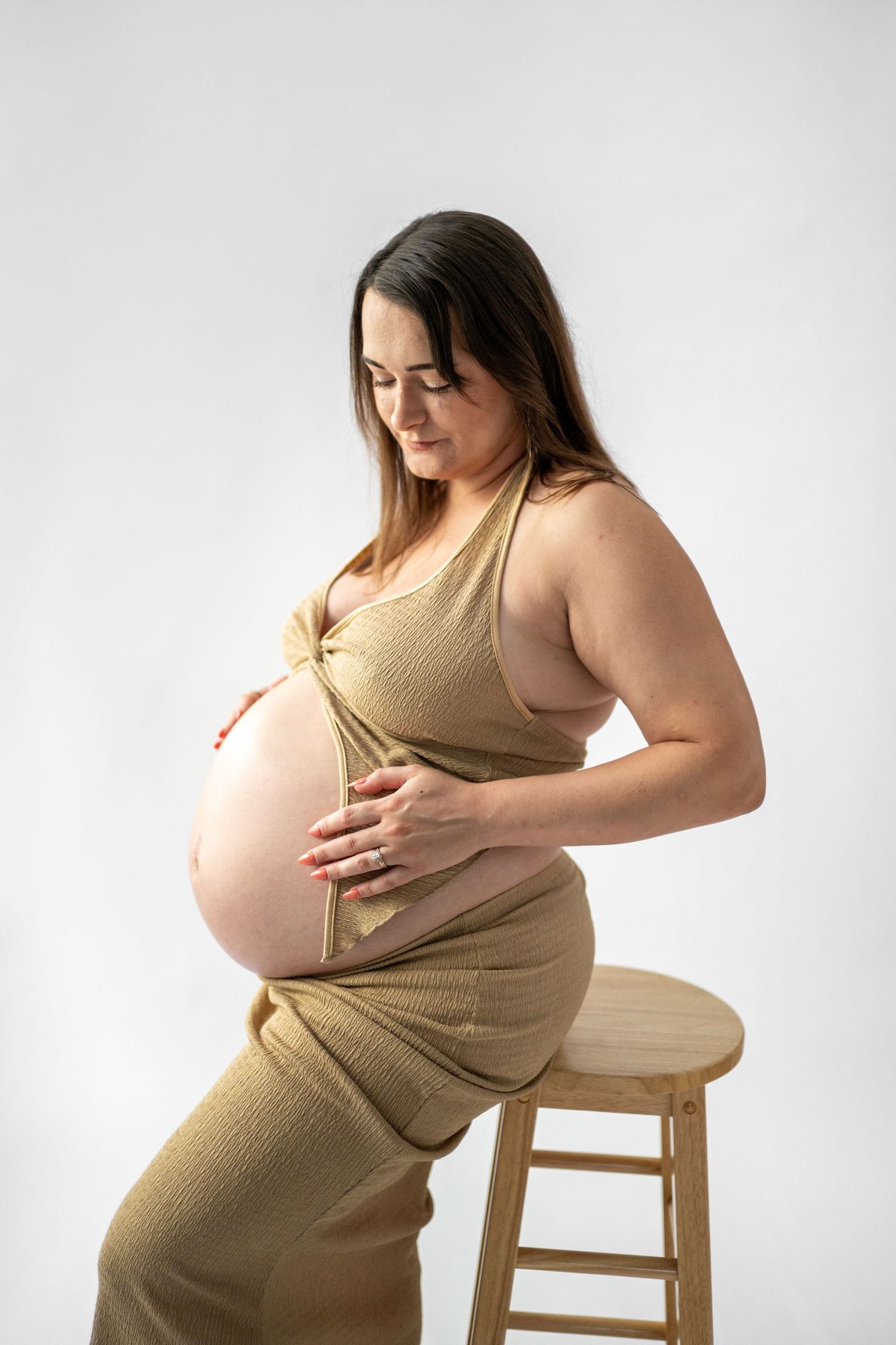 Maternity portrait series on wooden stool wearing beige dress.