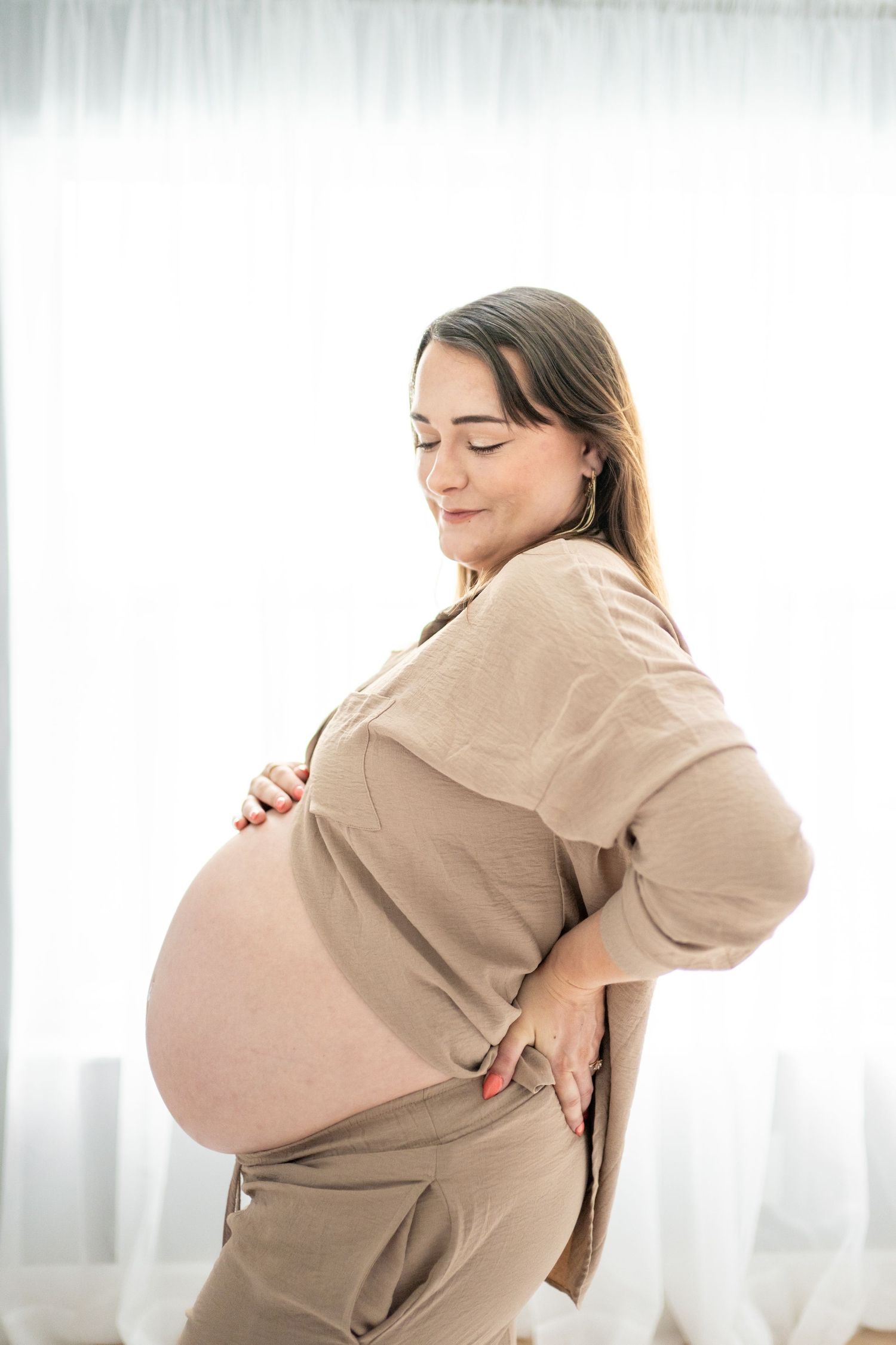 Pregnant woman in beige sweater holding her back while experiencing discomfort during pregnancy.