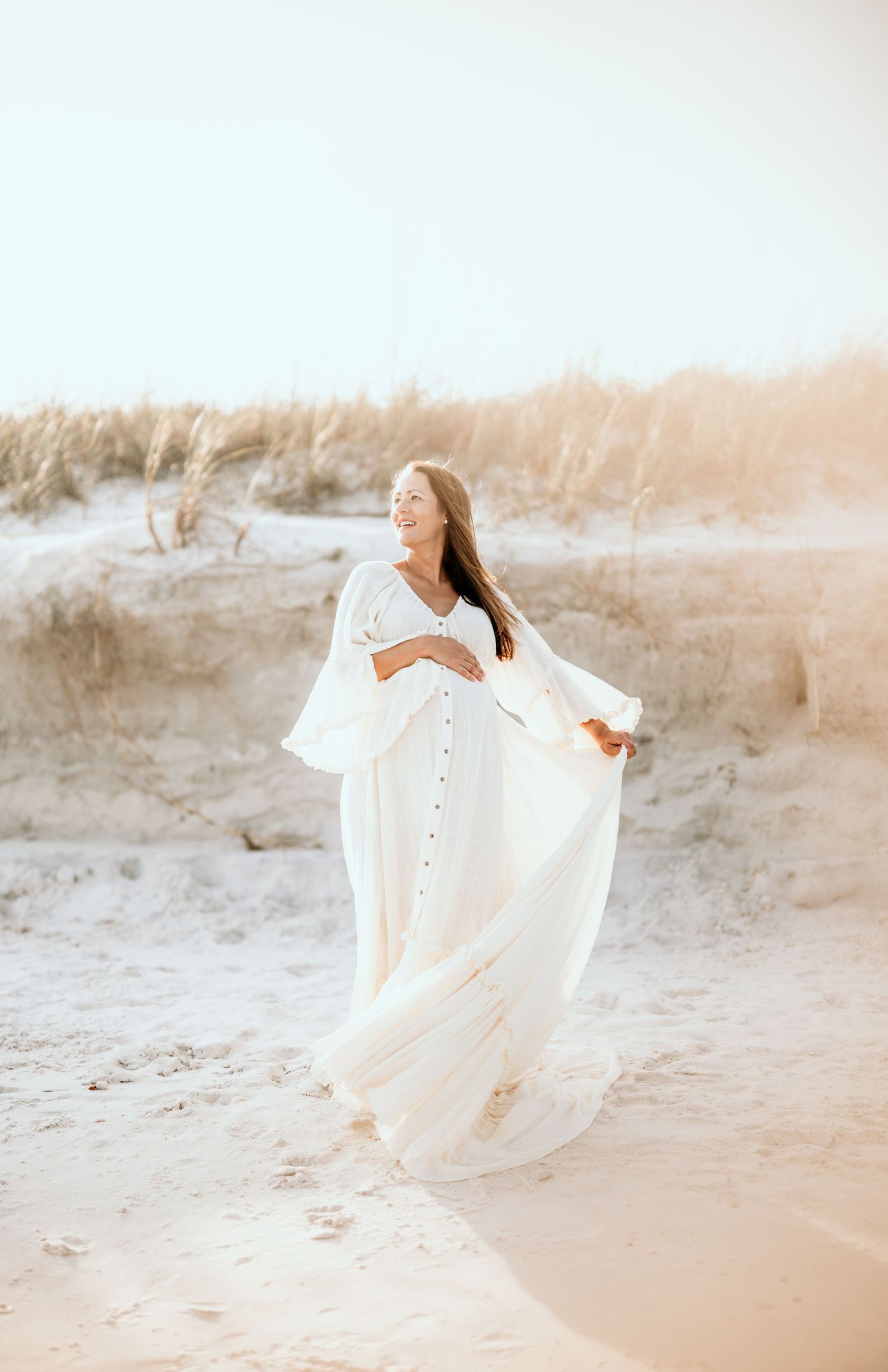 Dreamy maternity photo in white dress among sand dunes at golden hour.