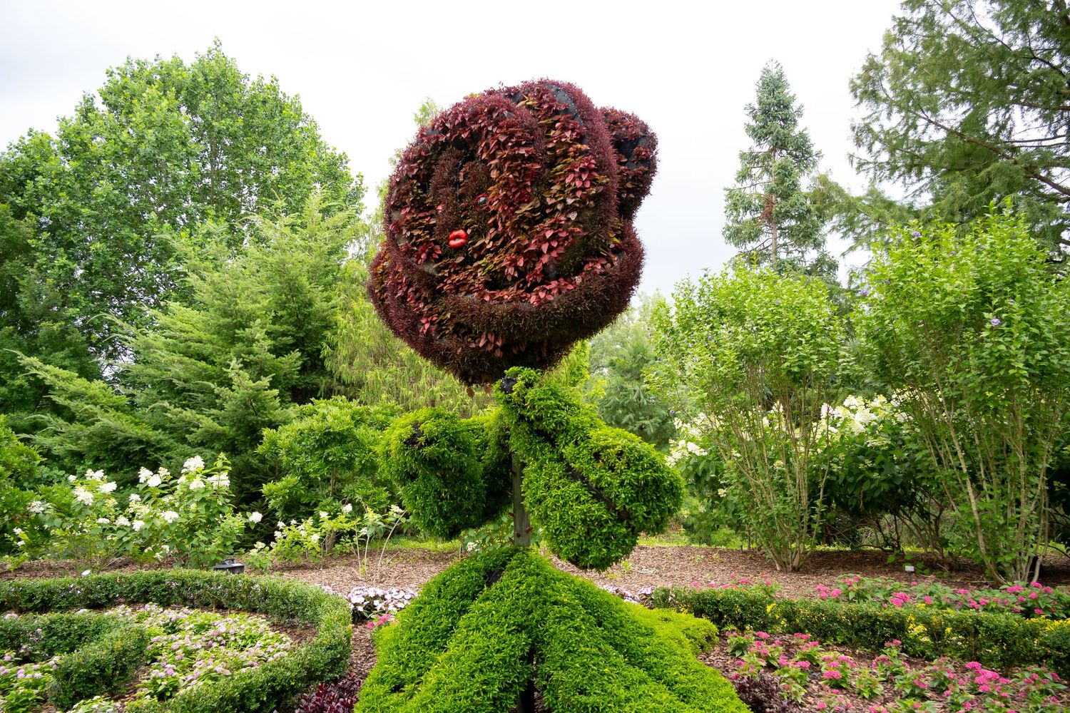 Large topiary sculpture with round burgundy flower head atop green shrub base in garden setting.