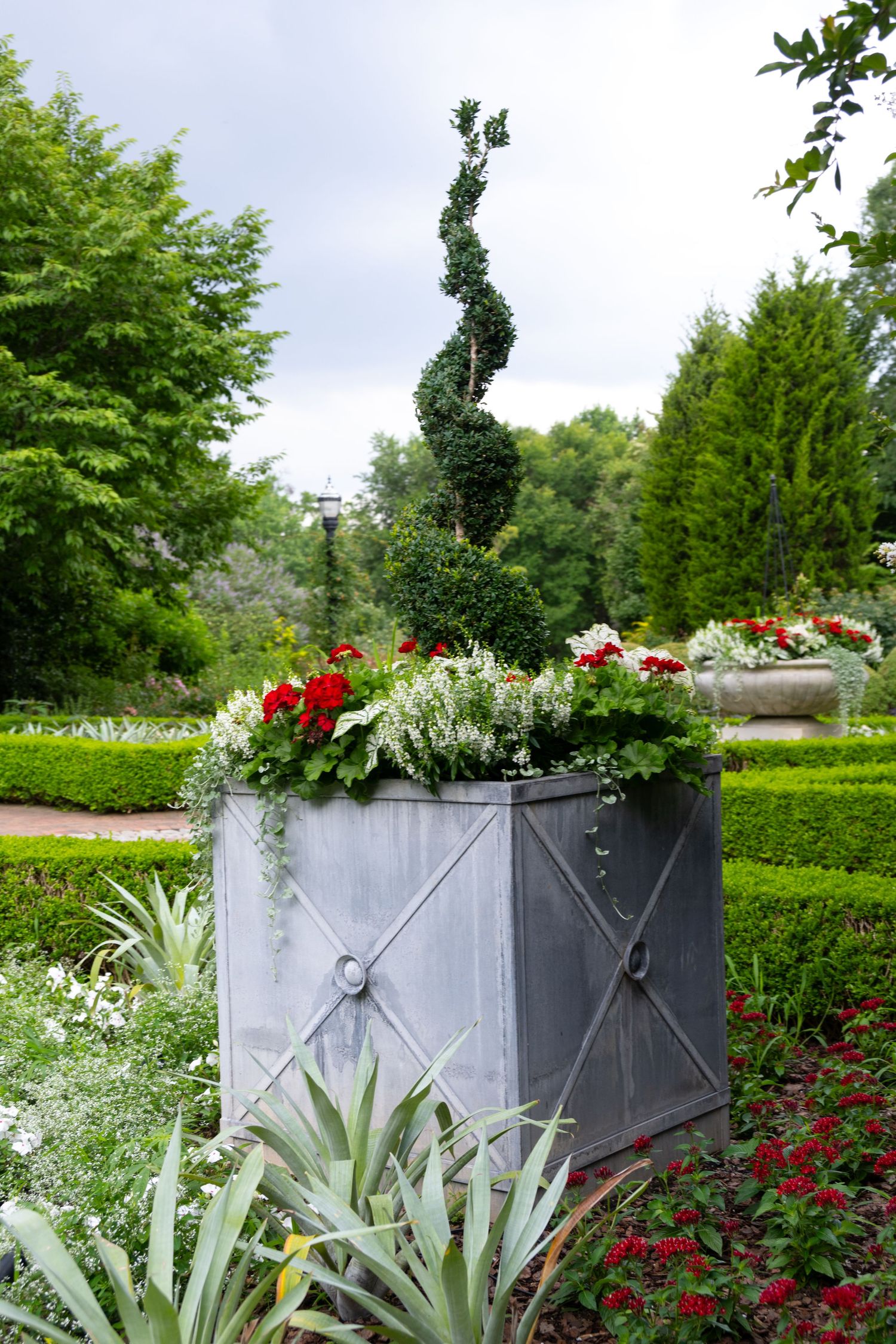Elegant metal planter with spiral topiary and colorful flowers in a formal garden setting.