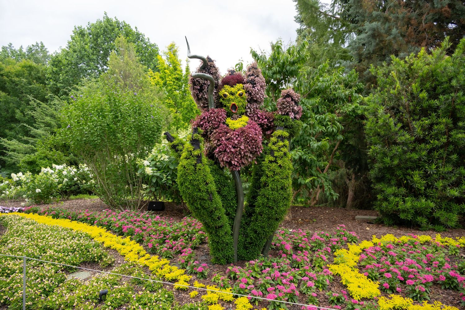 Artistic topiary sculpture with purple blooms surrounded by yellow border flowers in garden.