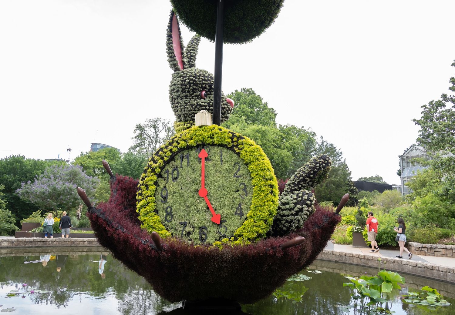Artistic topiary boat installation featuring a clock face and rabbit figure in a pond setting.