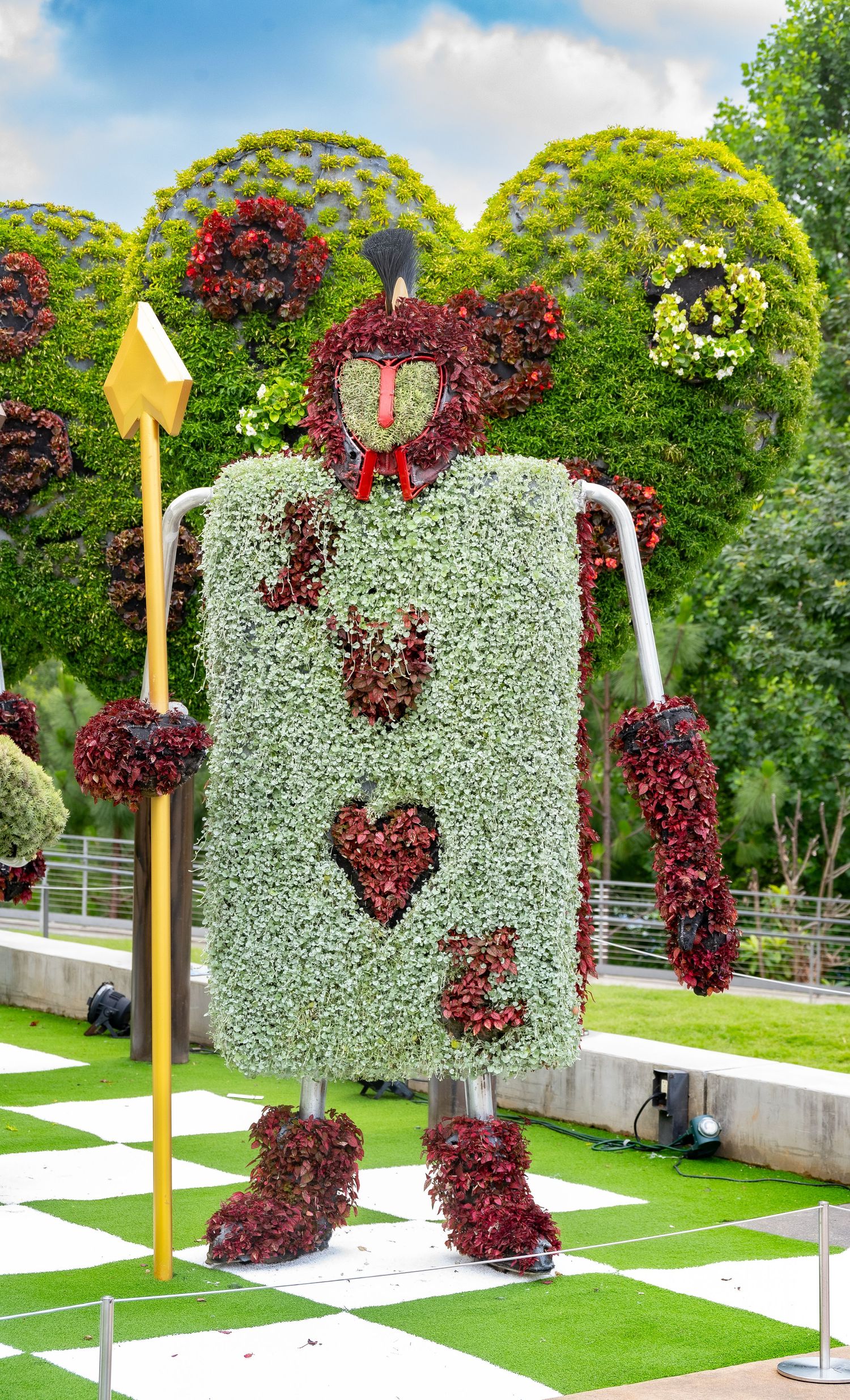 Whimsical garden topiary art sculpture of a playing card figure made with white and red flowers on a checkered lawn.