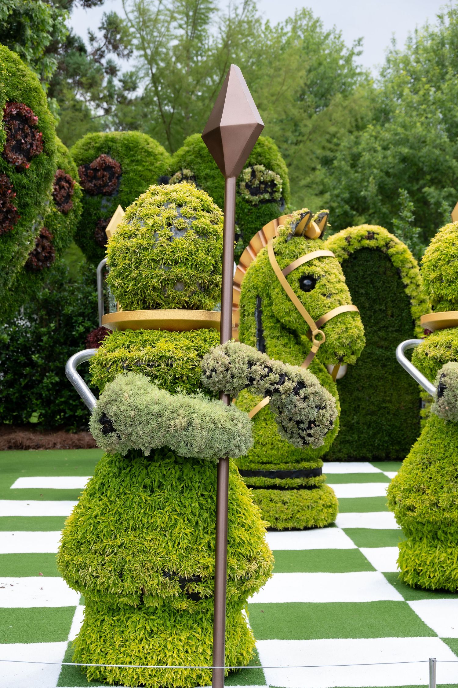 Garden topiary sculpture of a chess knight piece covered in green plants and flowers on a checkerboard pattern.
