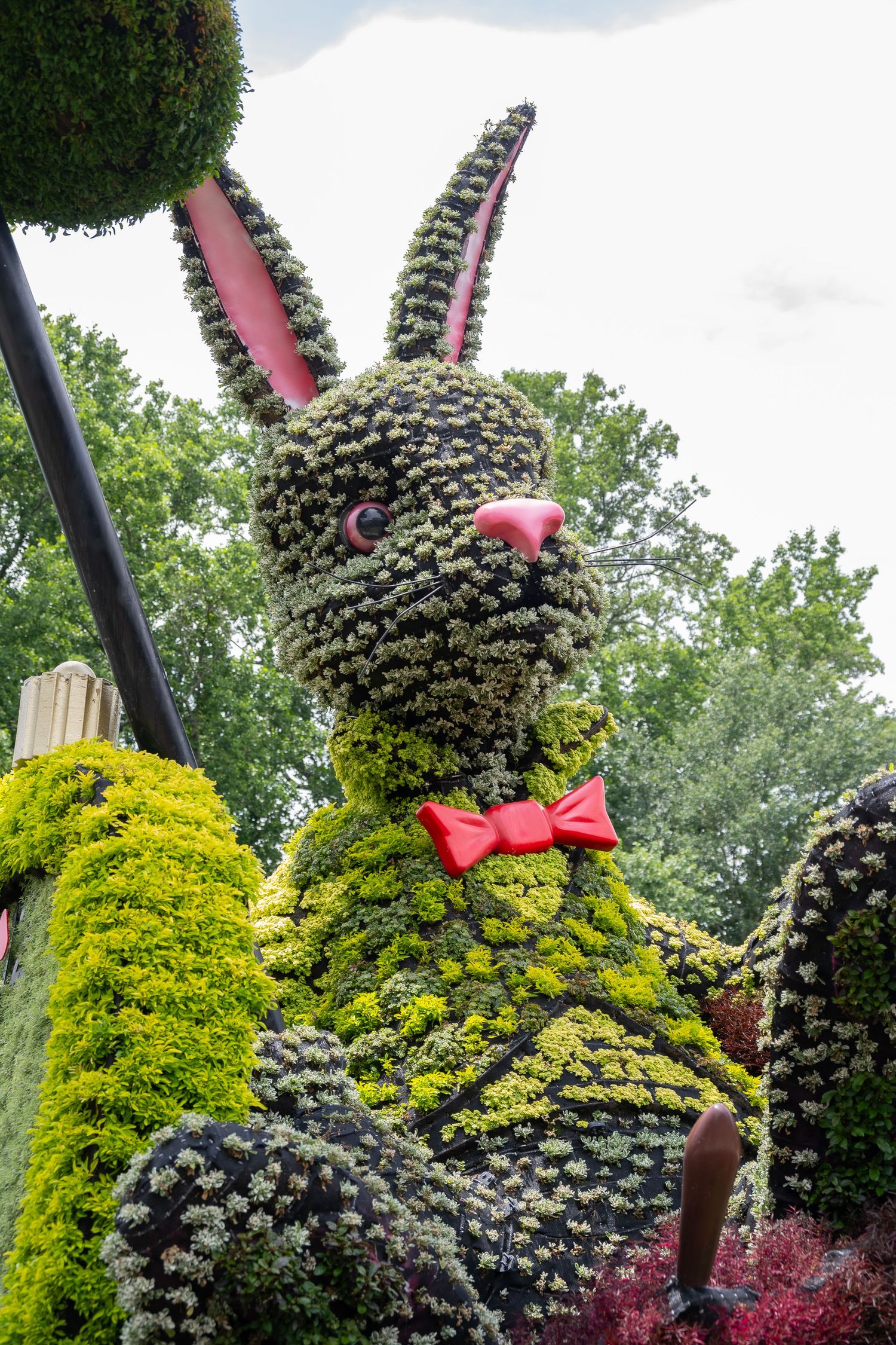 Large topiary rabbit sculpture with green foliage, pink accents and a red bowtie in a garden setting.