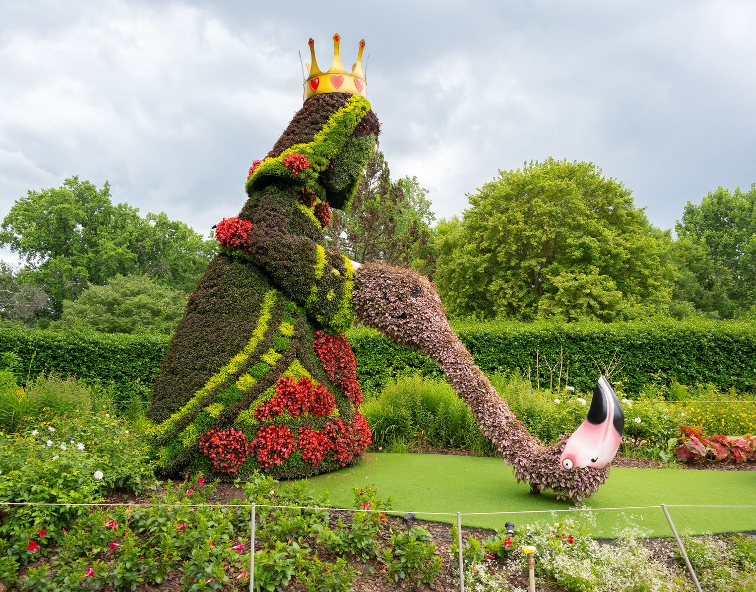 Whimsical garden topiary sculpture of a frog prince with crown on a green lawn surrounded by colorful flowers.