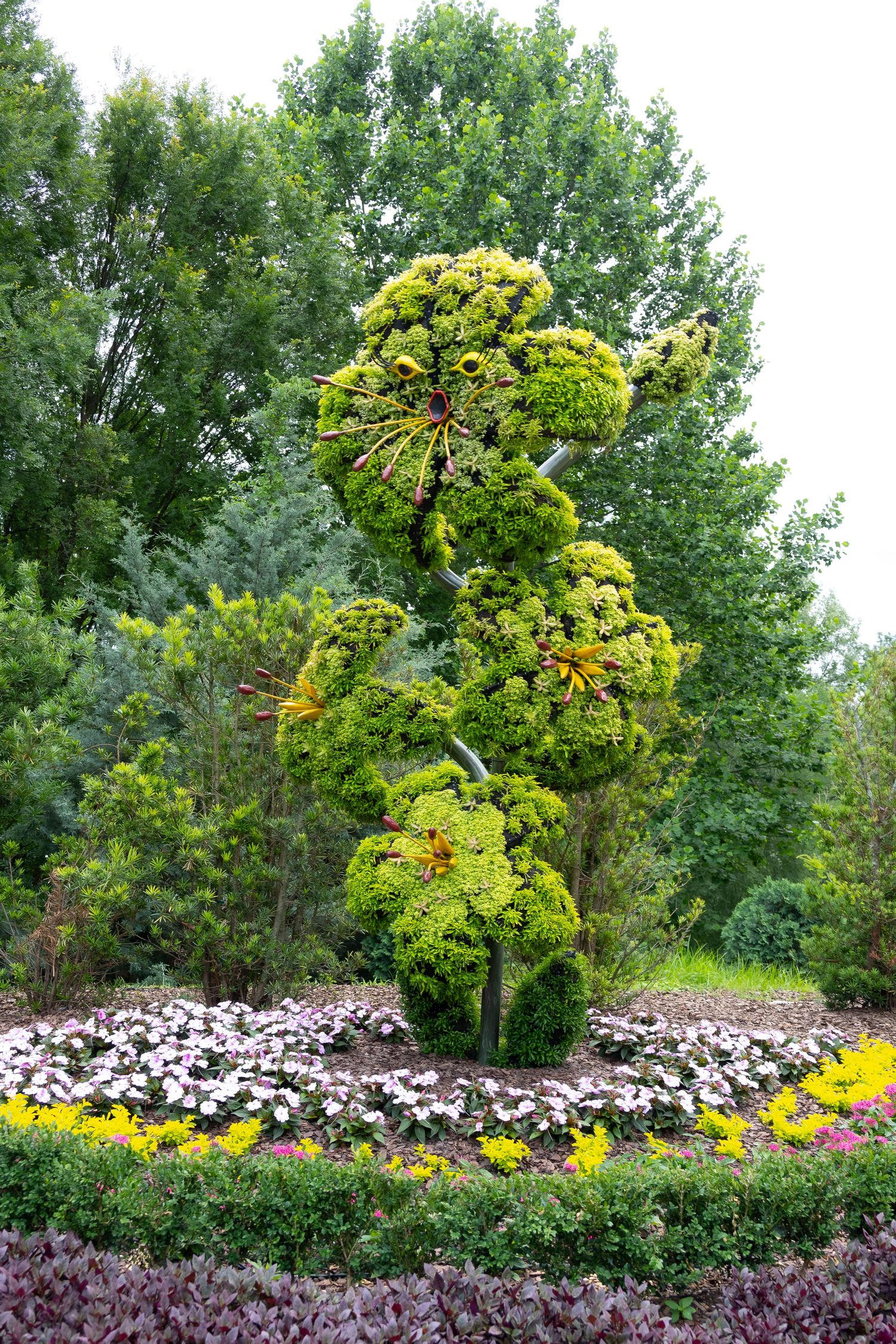 A tall, spiraling topiary sculpture surrounded by colorful flower beds in a garden setting.