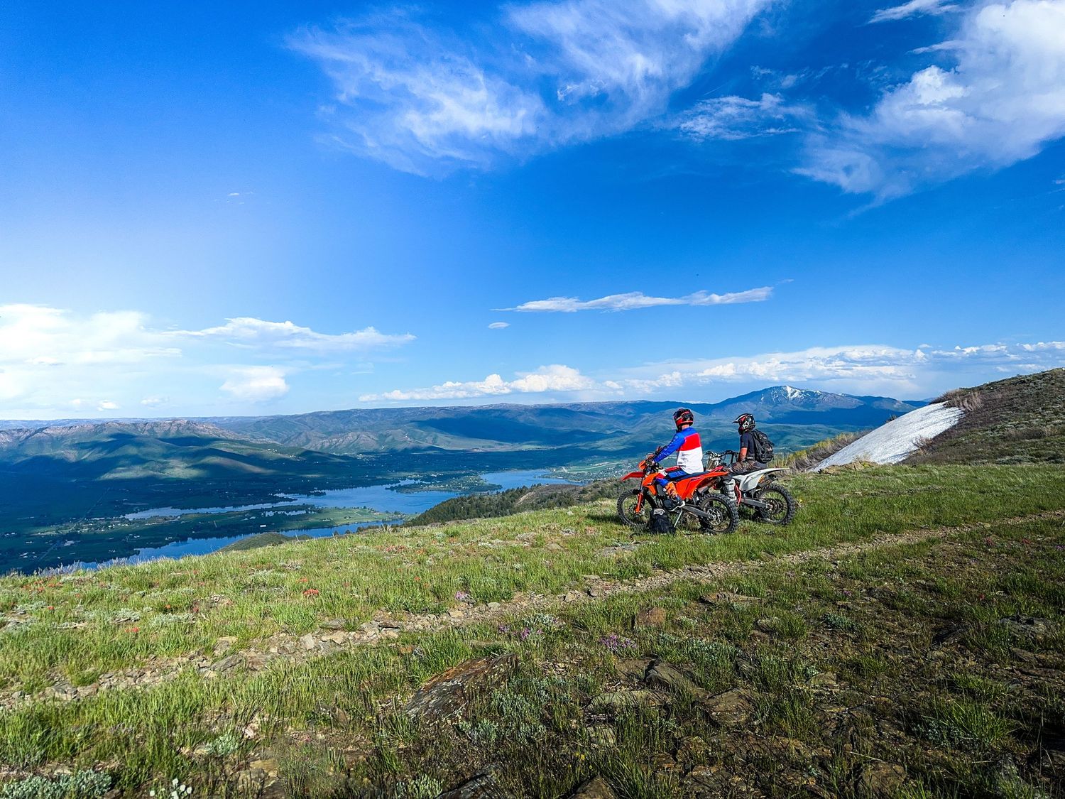 Two motocross riders pause on a lush green mountain slope overlooking a scenic lake and valley below.