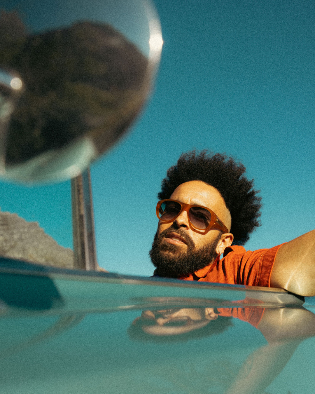 Eyewear brand advertisement showing person in sunglasses and orange shirt against vibrant turquoise background with PRAIA text.