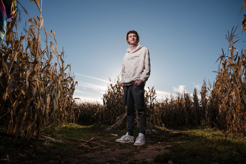 Full length photo of a high school senior boy in a white hoodie, jeans, and sneakers standing in a corn maze