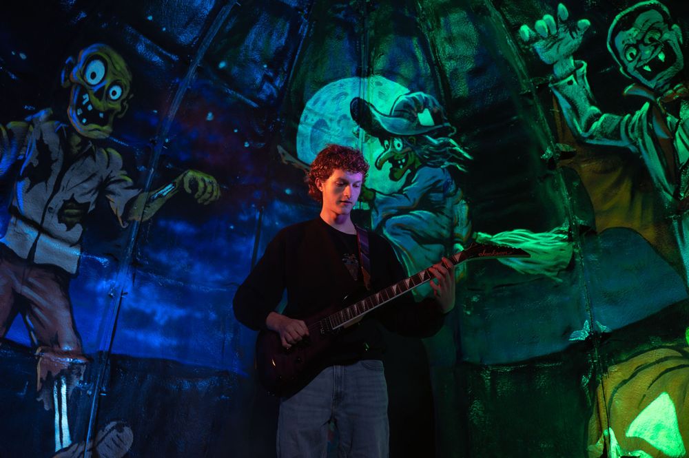 High school senior boy wearing a black sweatshirt playing guitar in a Halloween themed scene with a zombie, witch, and vampire