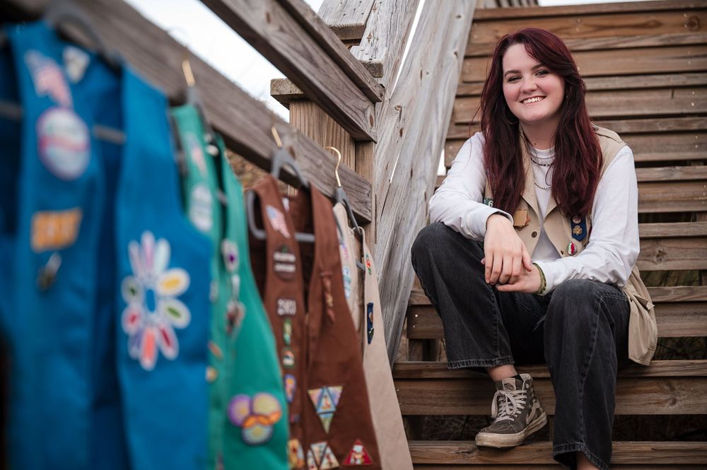 Portrait of a North Carolina high school senior with dark hair and glasses sitting on sand dune stairs with her Girl Scouts of America vests
