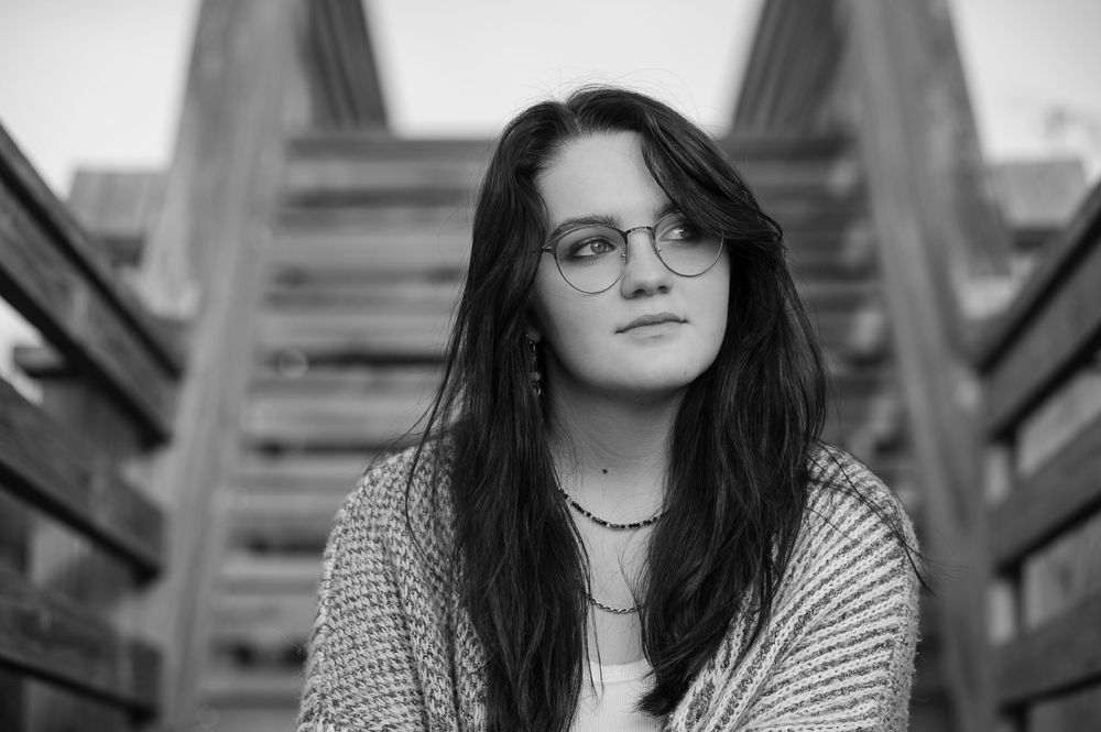 Black and white portrait of a North Carolina high school senior with dark hair and glasses sitting on sand dune stairs in Nagsa Head, NC