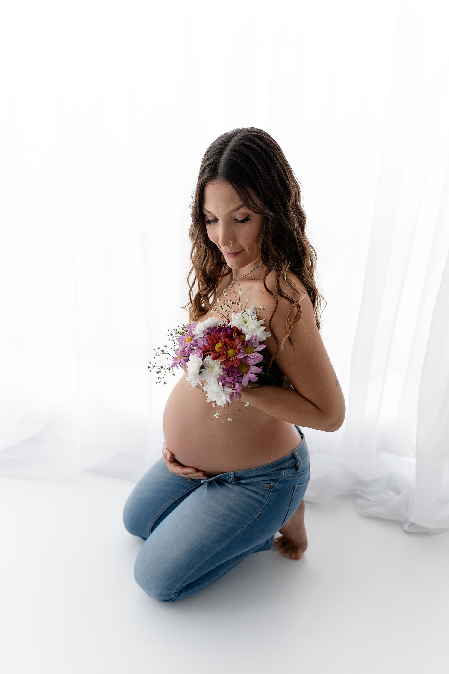 Pregnant woman kneels while holding fresh flowers against her belly in white studio setting.