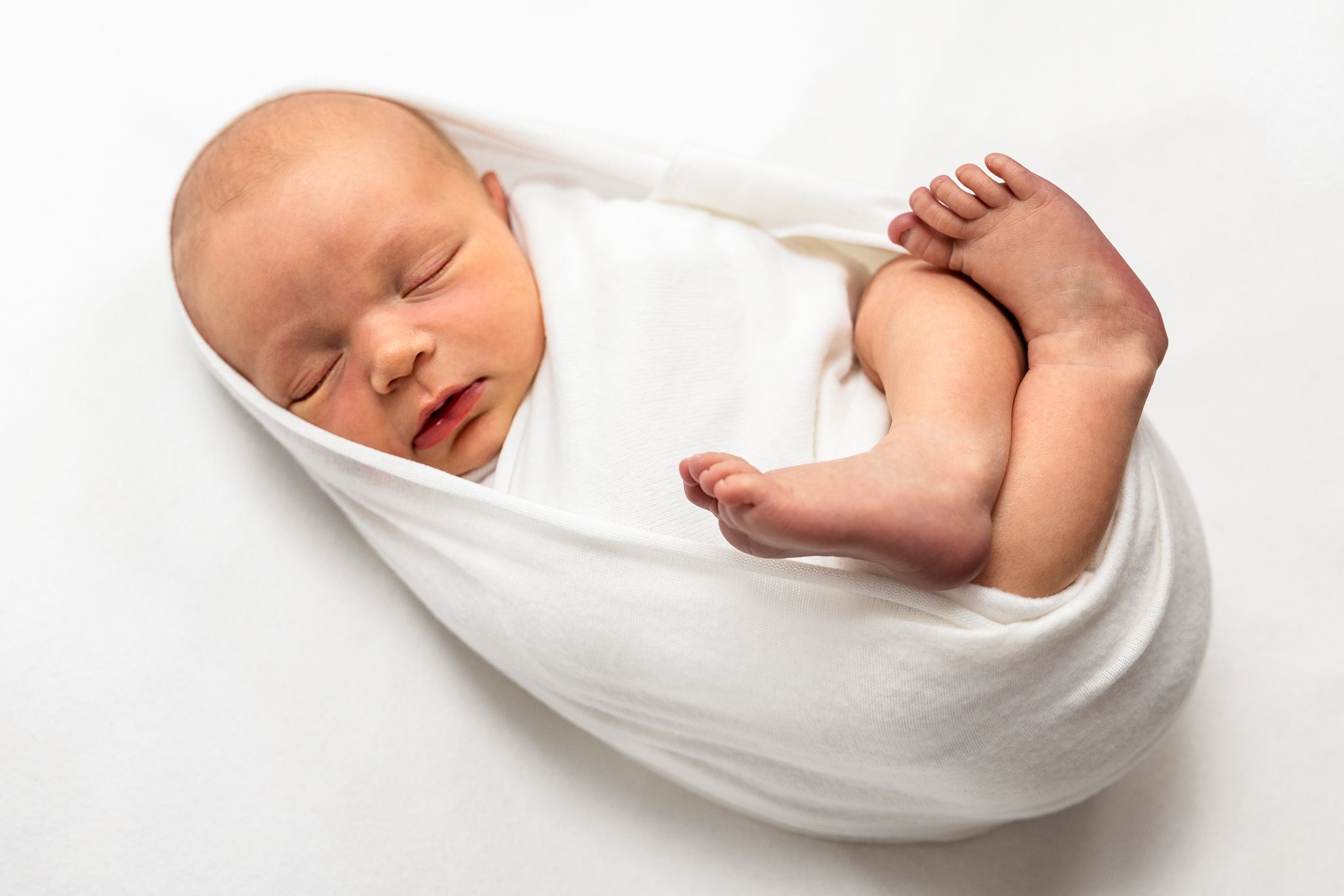 A peaceful sleeping newborn baby wrapped in soft white fabric against a white background.