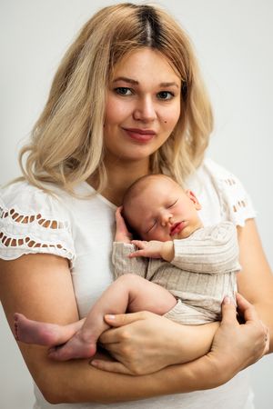 A mother in a white lace top holds her sleeping newborn baby wrapped in a beige blanket.