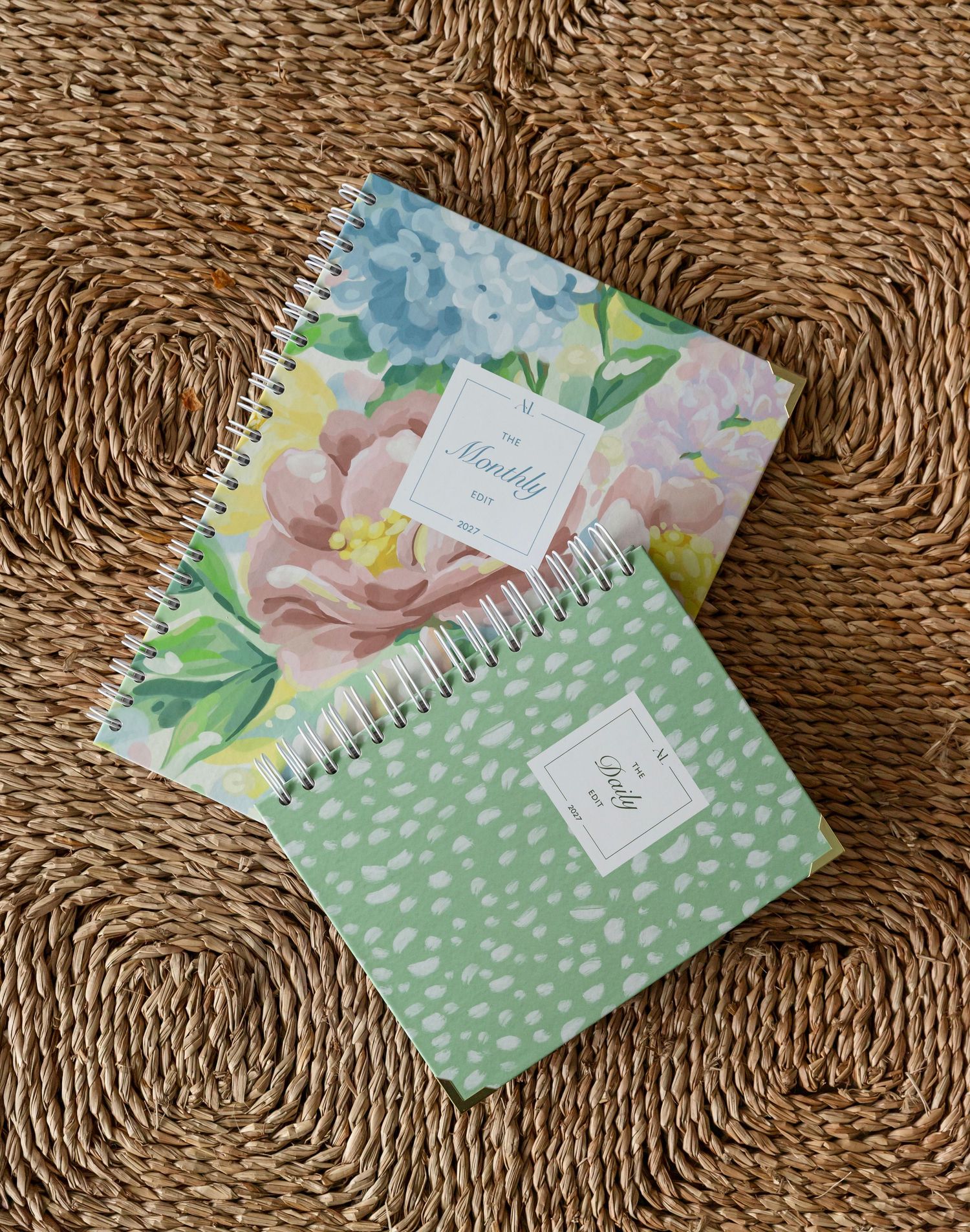 Two spiral-bound planners with floral and green dotted covers rest on a woven rattan surface.